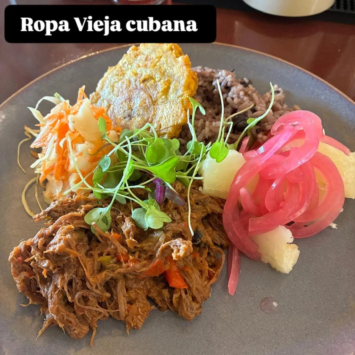A plate of traditional Cuban food including shredded beef, rice and beans, cooked vegetables, mashed potatoes, pickled red onions, and microgreens, with a label stating "Ropa Vieja cubana."