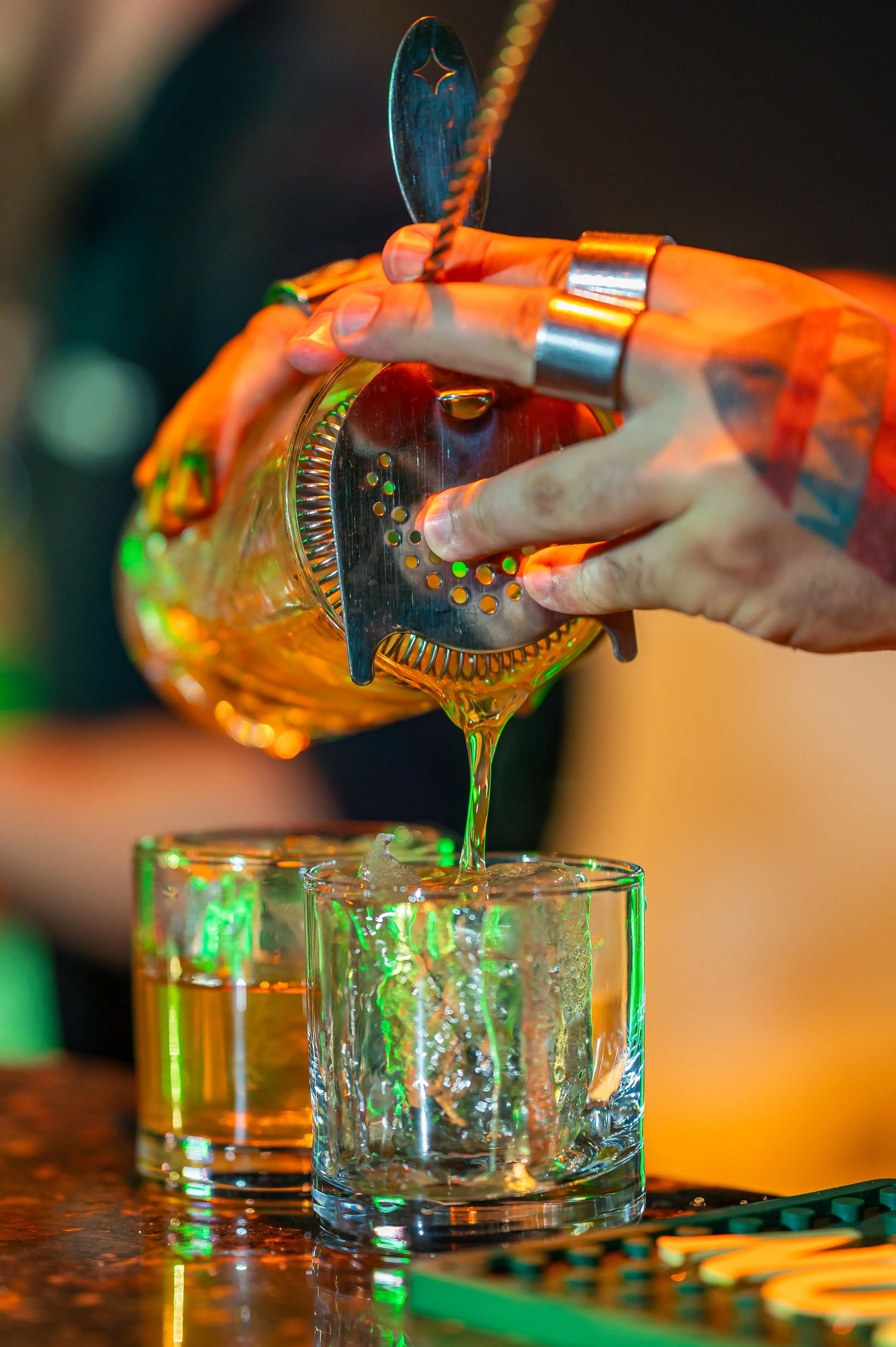 A person with tattoos on their hand is pouring a drink over ice in a glass, using a metal cocktail strainer.