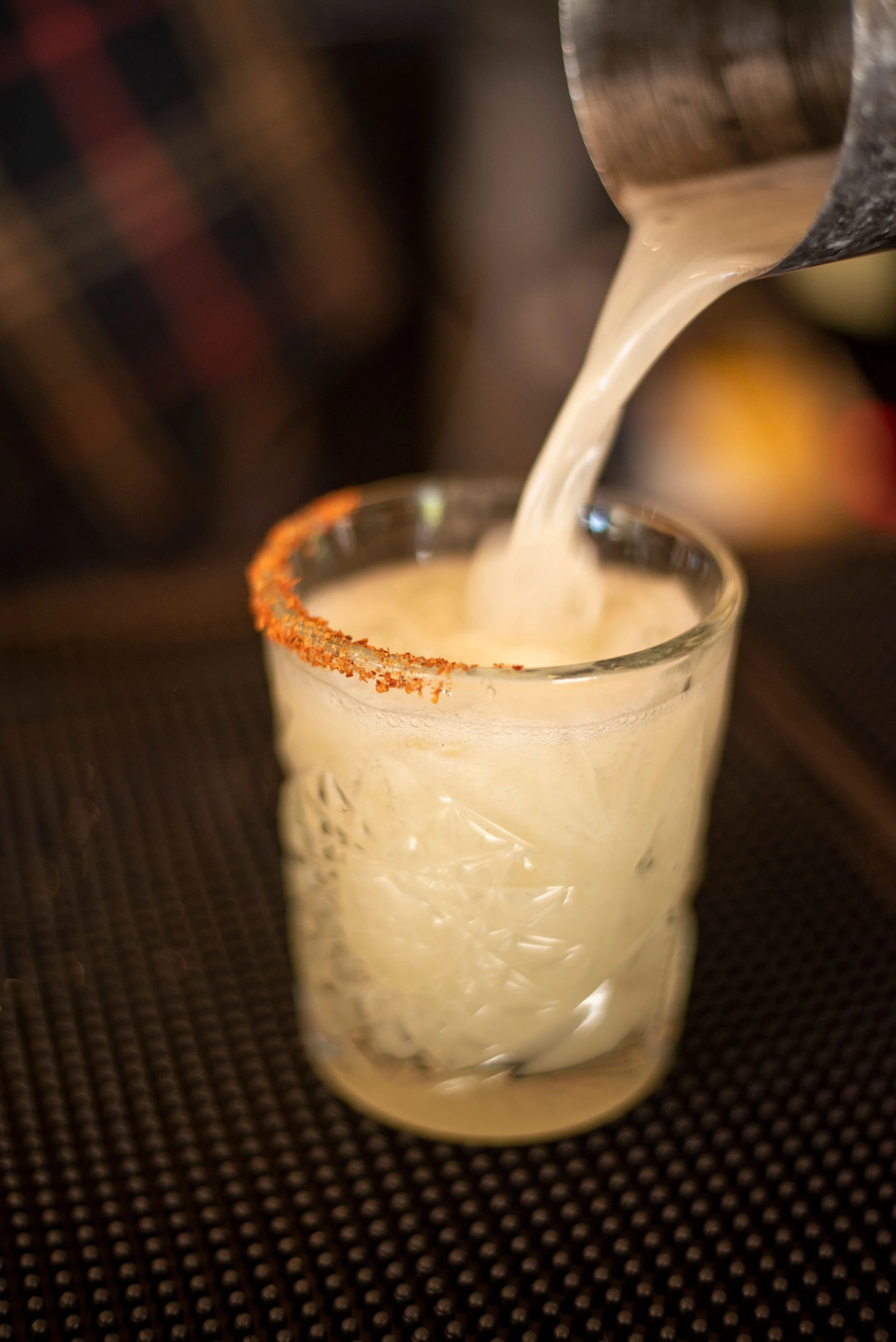 Close-up of a glass with a salted rim being filled with a creamy beverage, possibly a cocktail, on a dark textured surface.