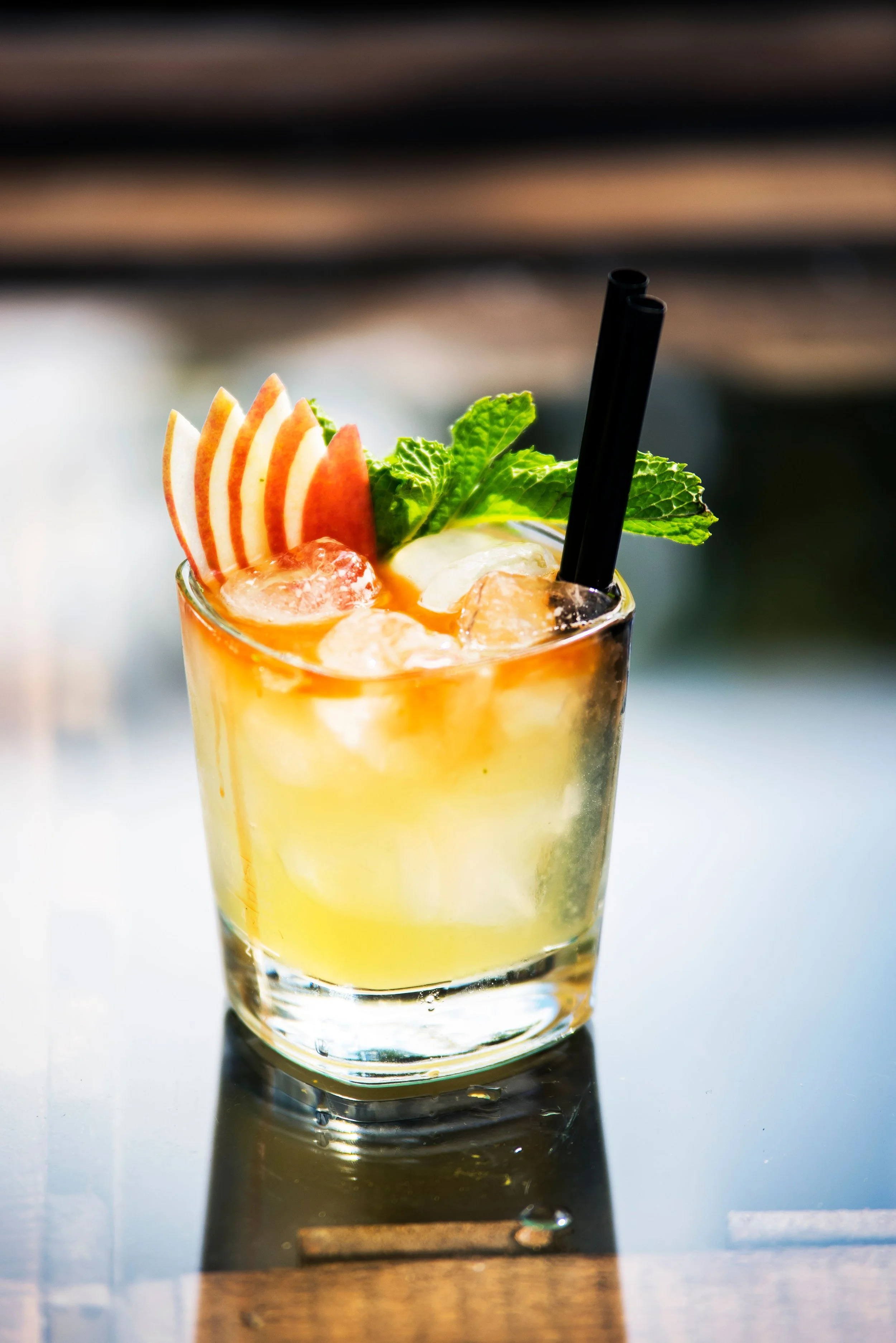 A cocktail drink in a glass, garnished with apple slices, fresh mint leaves, a strawberry, and two black straws, placed on a dark reflective surface.