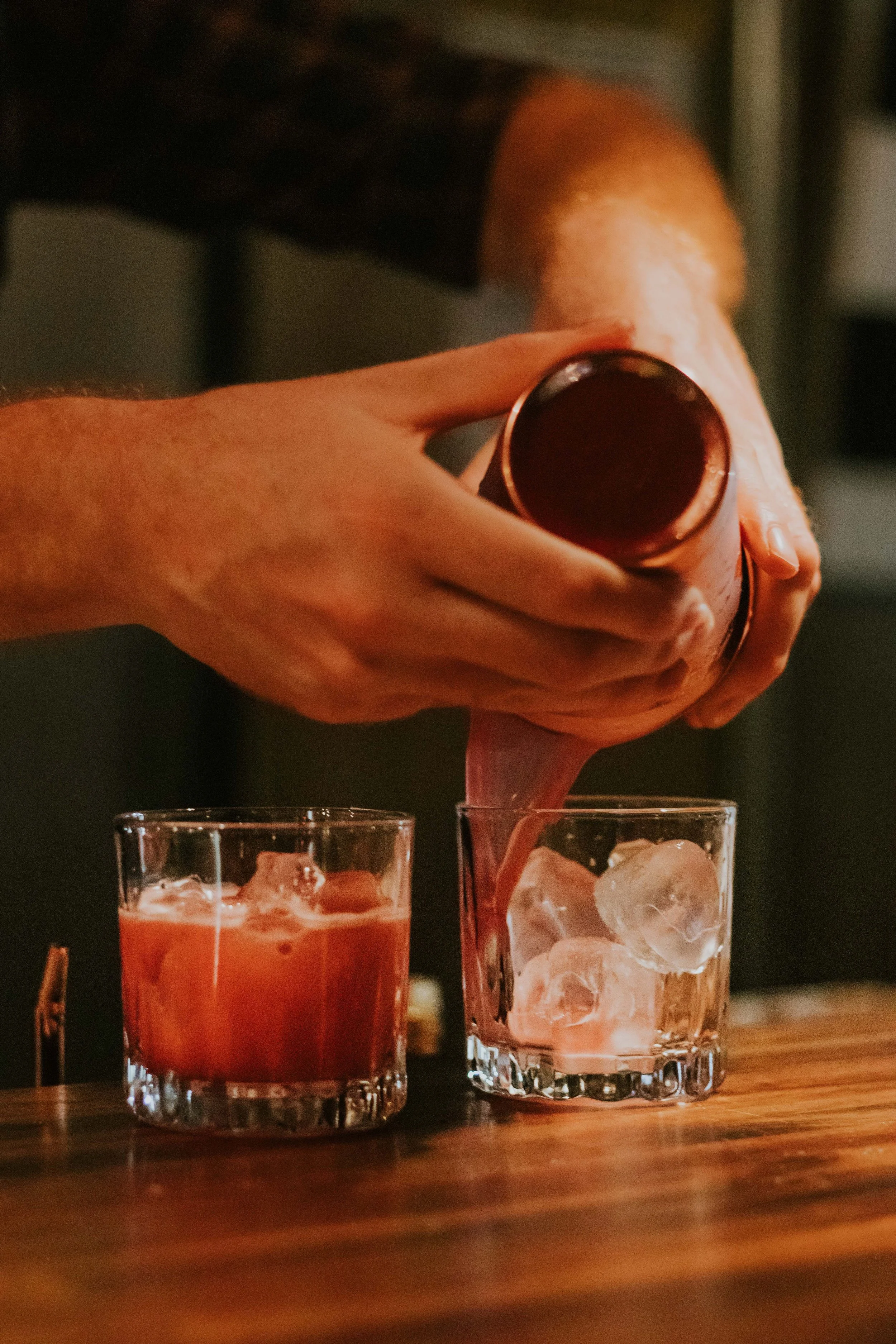 A person pouring a dark beverage into a glass filled with ice, with two glasses of drinks on a wooden table in a dimly lit setting.