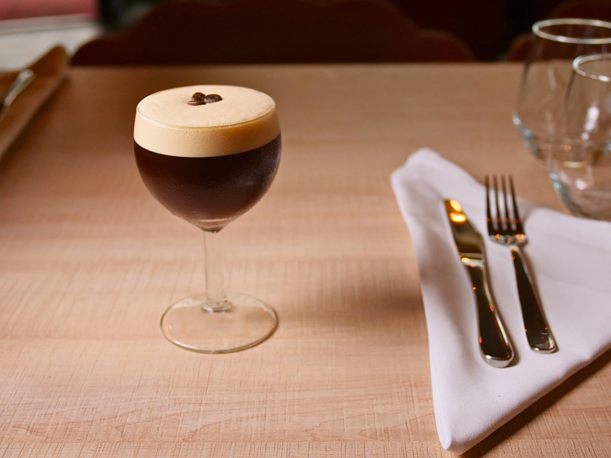 A glass of dark beer with a frothy head garnished with coffee beans sits on a wooden table next to a white cloth napkin and a set of silverware, with empty drinking glasses in the background.