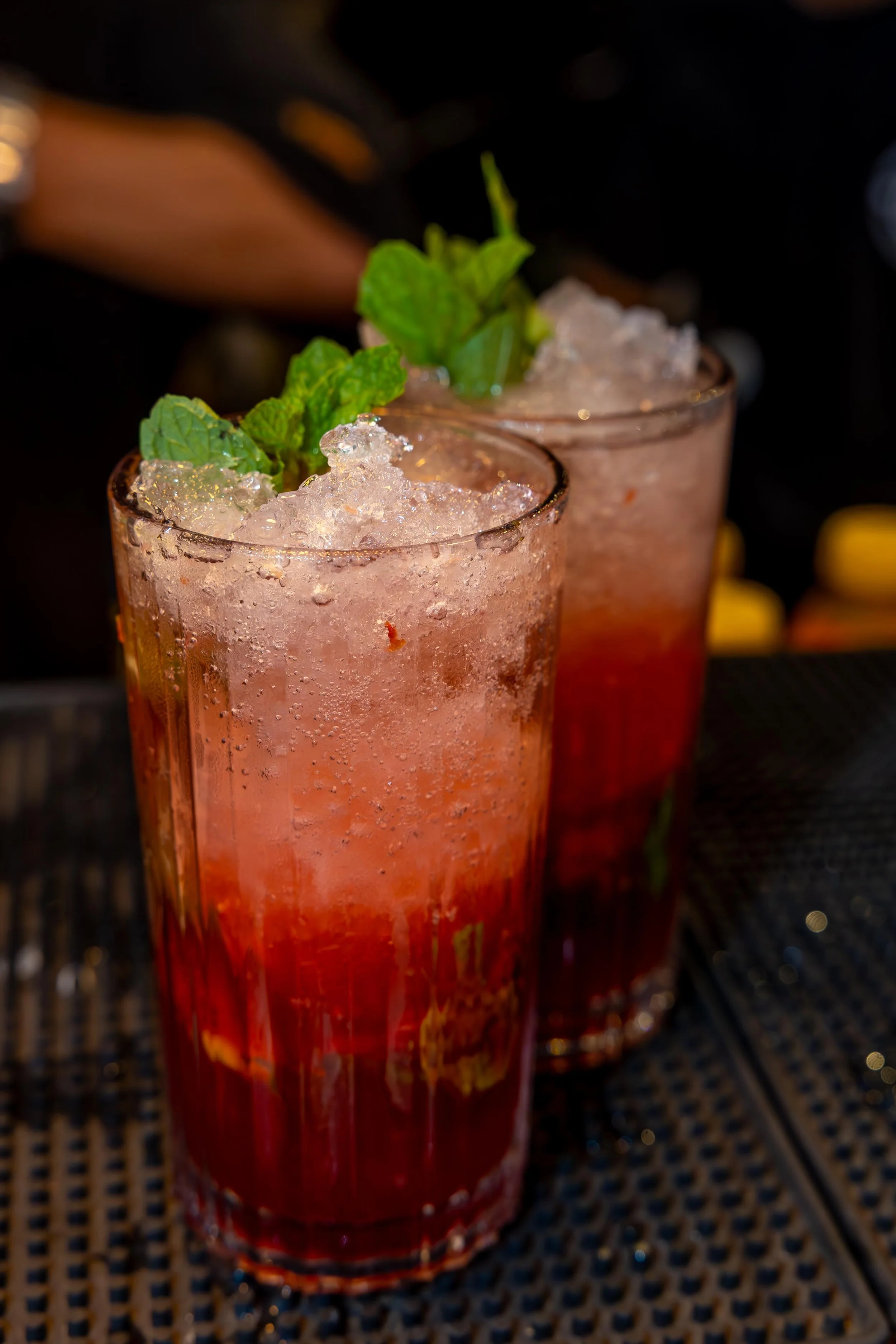 Two tall glasses filled with a pink beverage topped with crushed ice, garnished with fresh mint leaves, on a dark woven surface background.