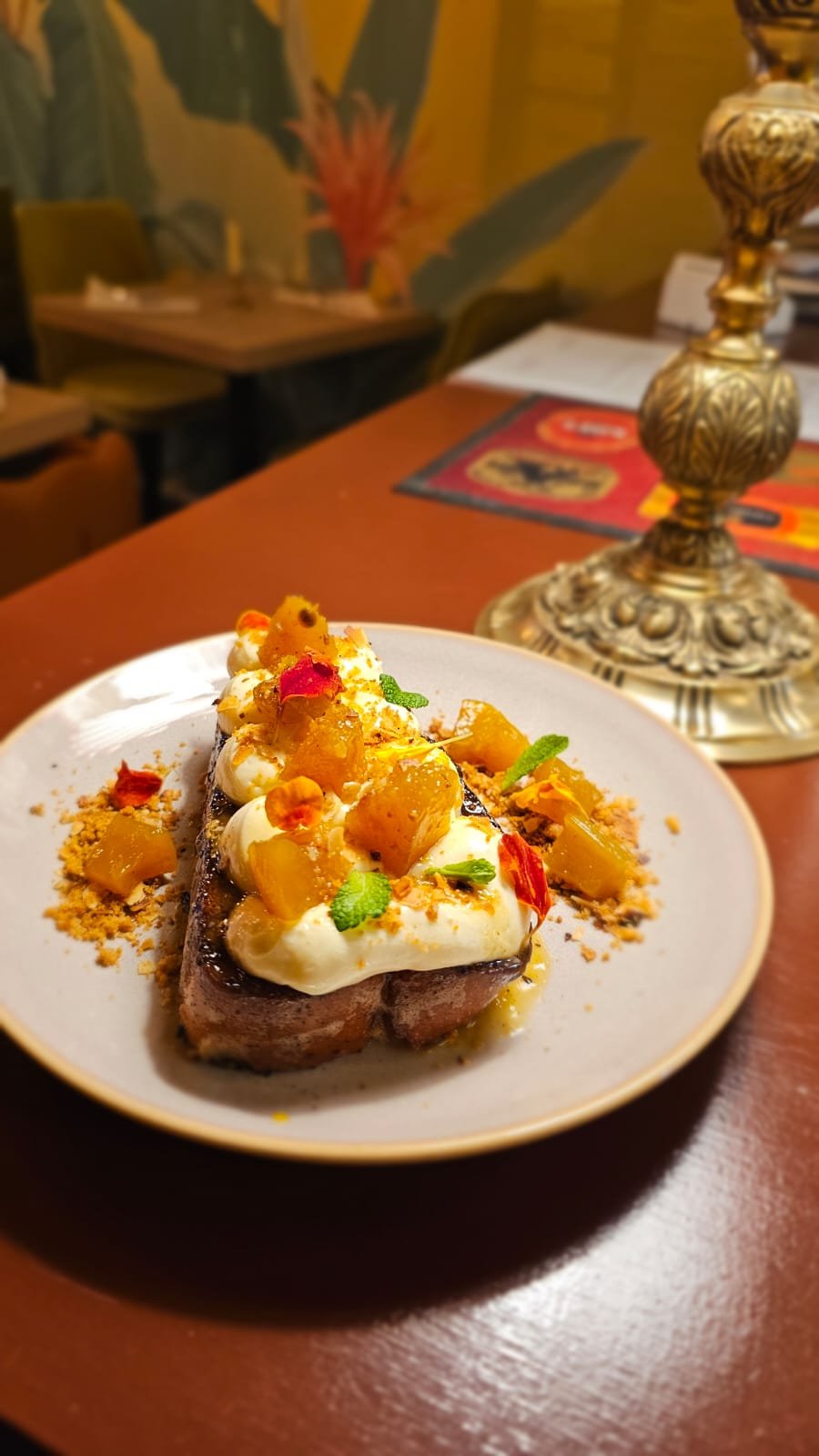 A plated dessert brioche with a slice of cake topped with whipped cream, pineapple chunks, edible flowers, and garnished with green herbs, served on a white plate in a restaurant setting.