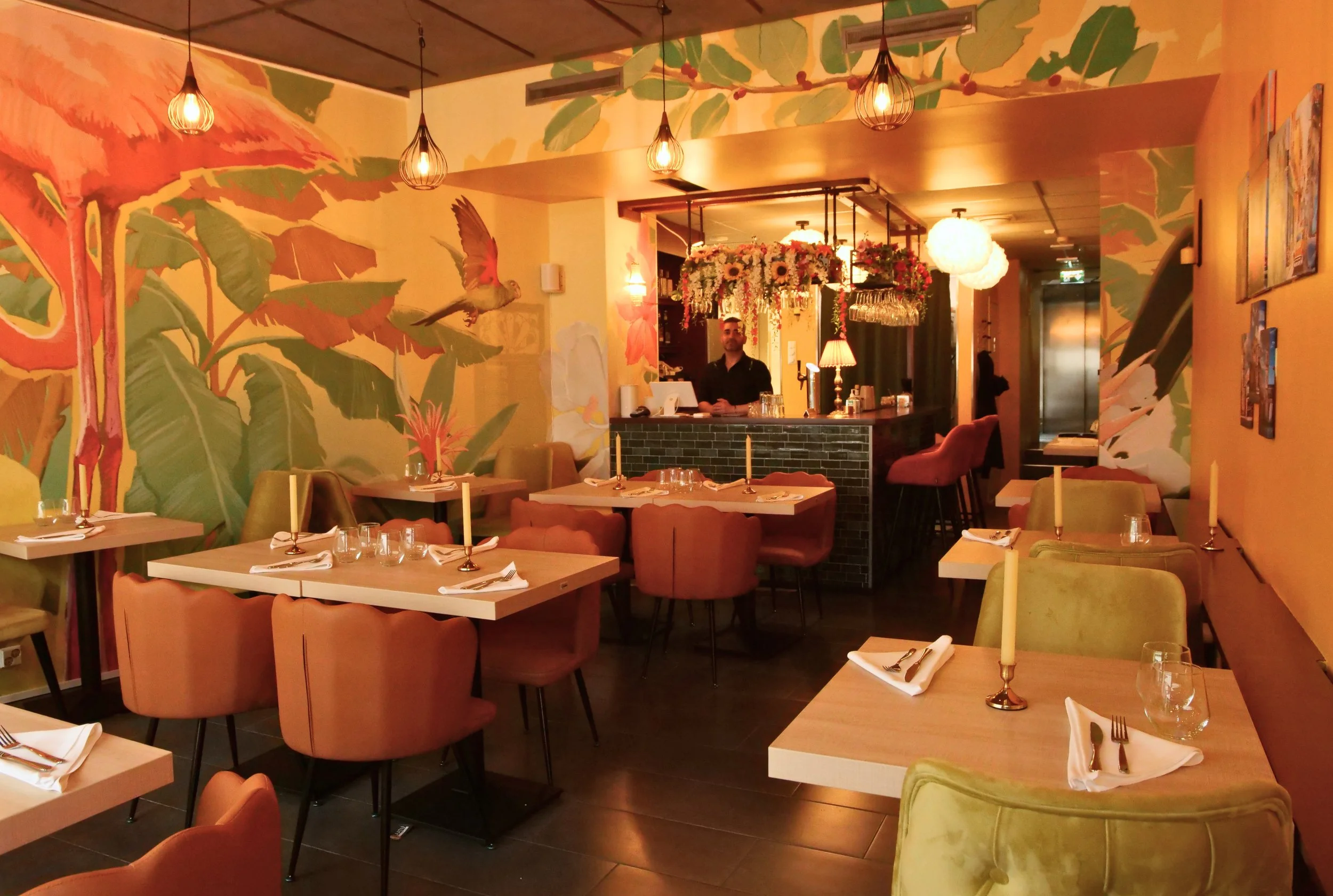 Interior of a restaurant with decorated walls featuring large banana leaves, a hummingbird, and flowers. There are tables set with candles, glasses, and napkins, and a bartender standing behind the bar adorned with hanging flower arrangements and wine glasses.