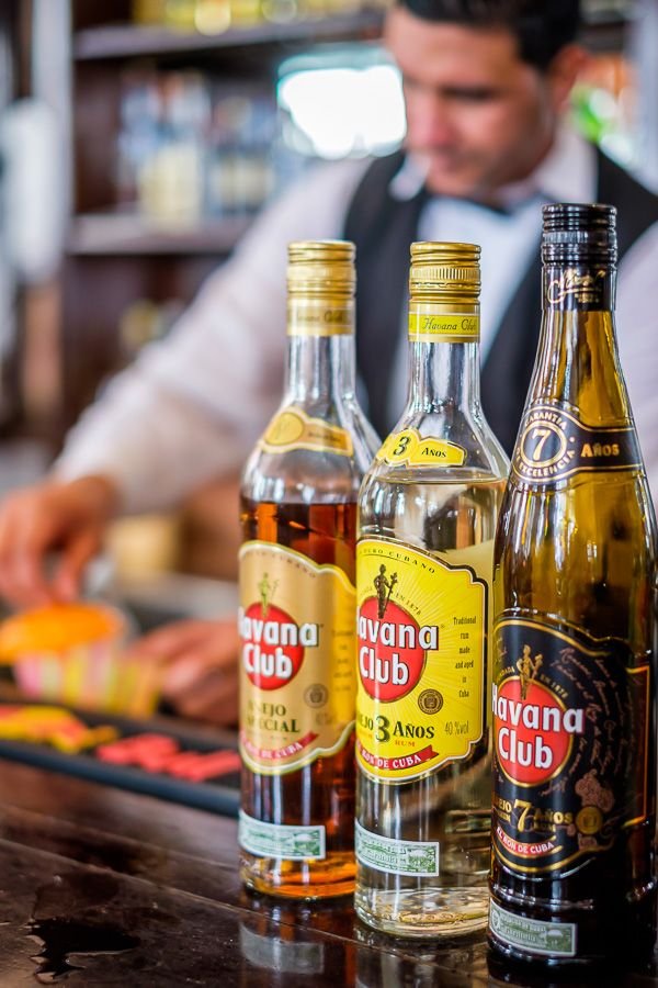 Three bottles of Havana Club rum on a bar counter with a bartender in the background.