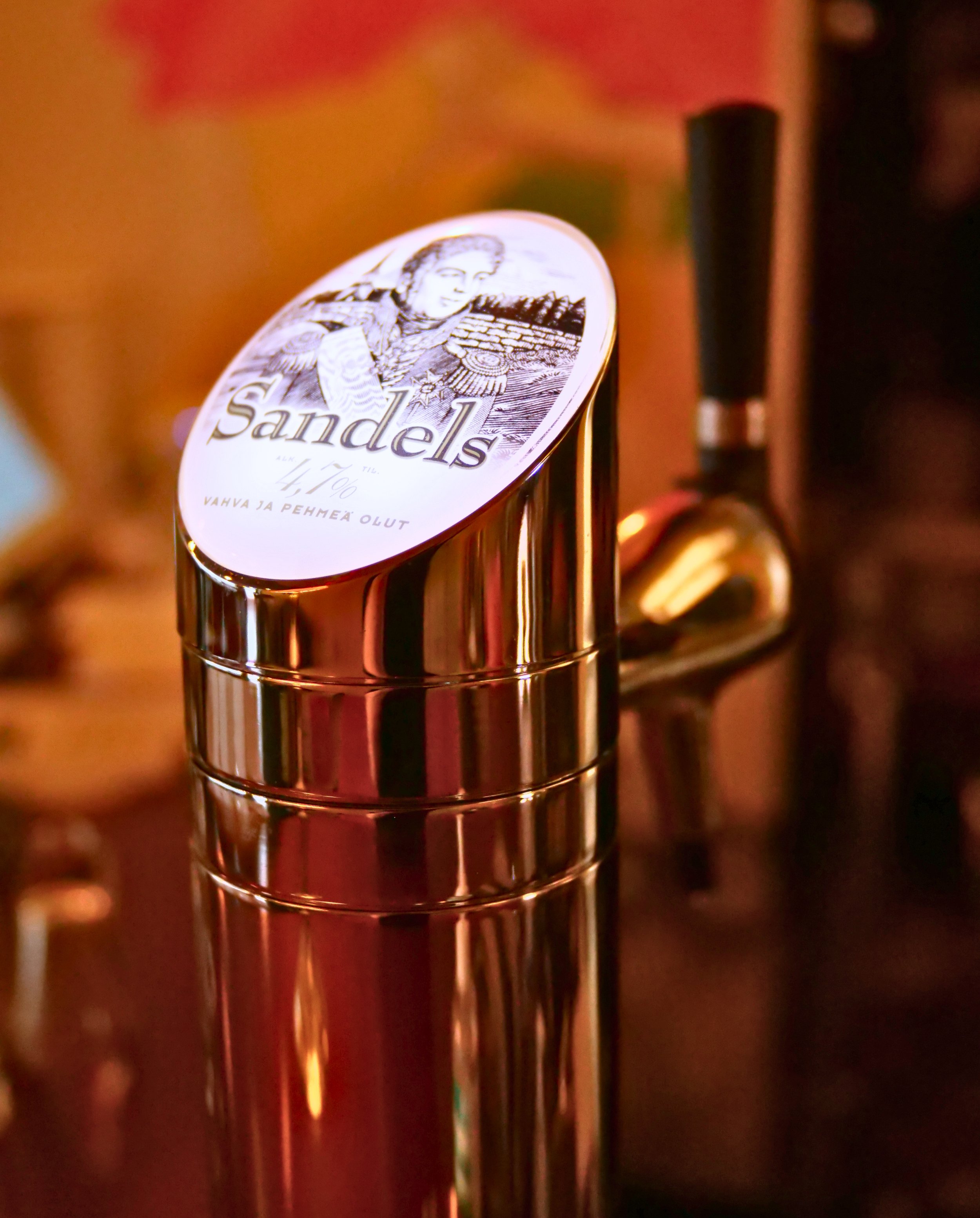 Close-up of a beer tap handle with a label reading 'Sandels', depicting a woman in traditional attire, set against a warm, indoor background.