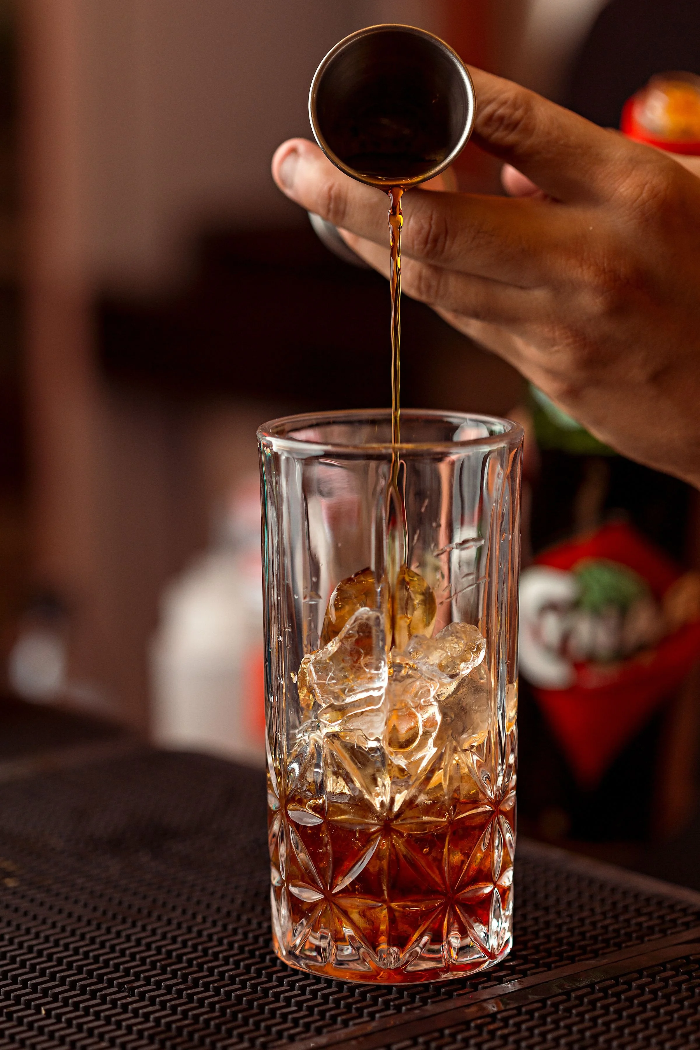 A close-up of a hand pouring dark liquid, likely coffee, from a small metal container into a tall glass filled with ice cubes.