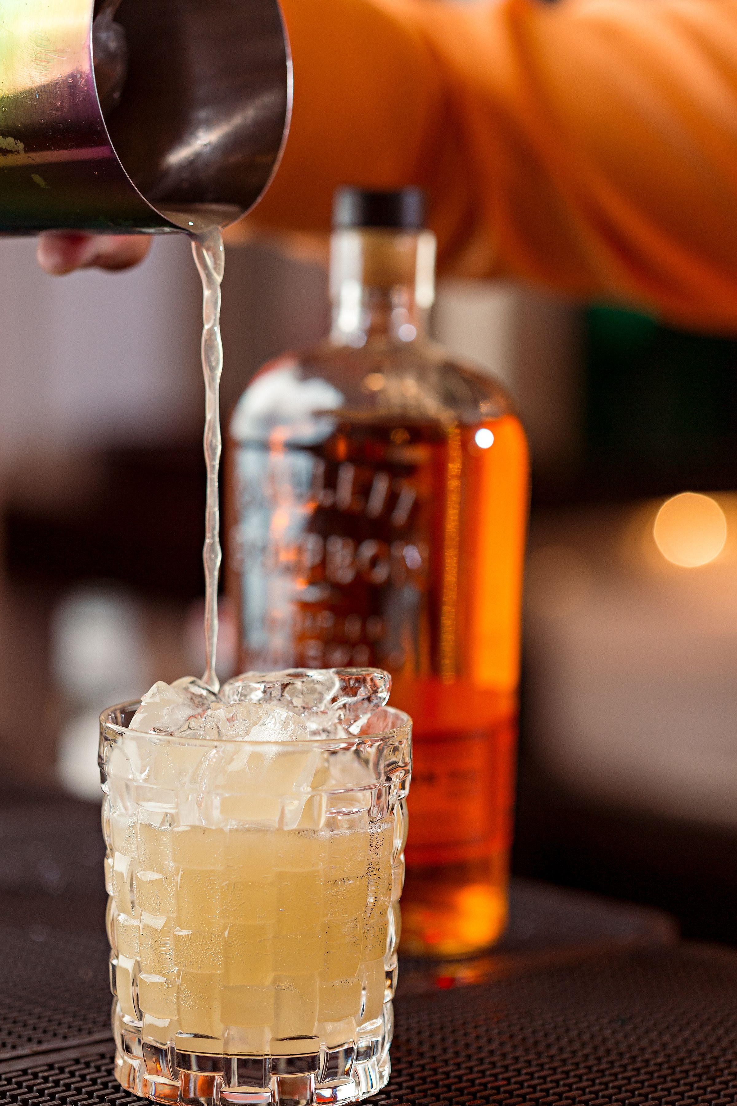 A glass of light-colored drink with ice cubes, with liquid being poured into it from a metal shaker, and a bottle of alcohol in the background.