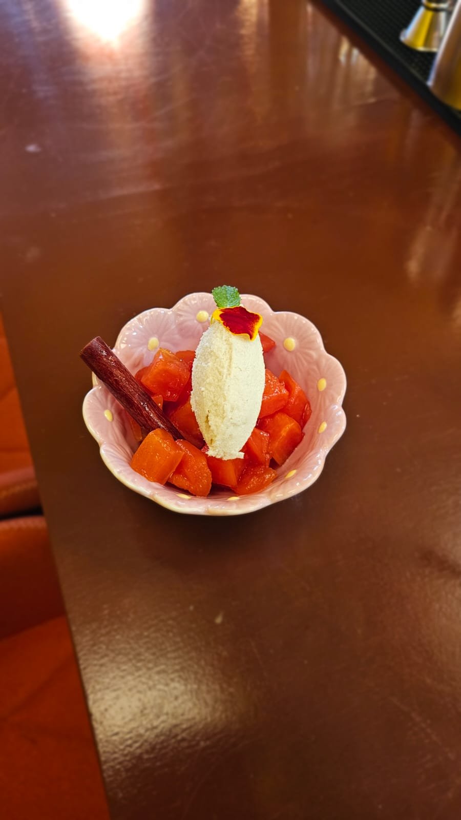 Dessert with diced orange fruit, a scoop of vanilla ice cream, a cinnamon stick, and decorative garnish in a pink floral bowl on a wooden table.
