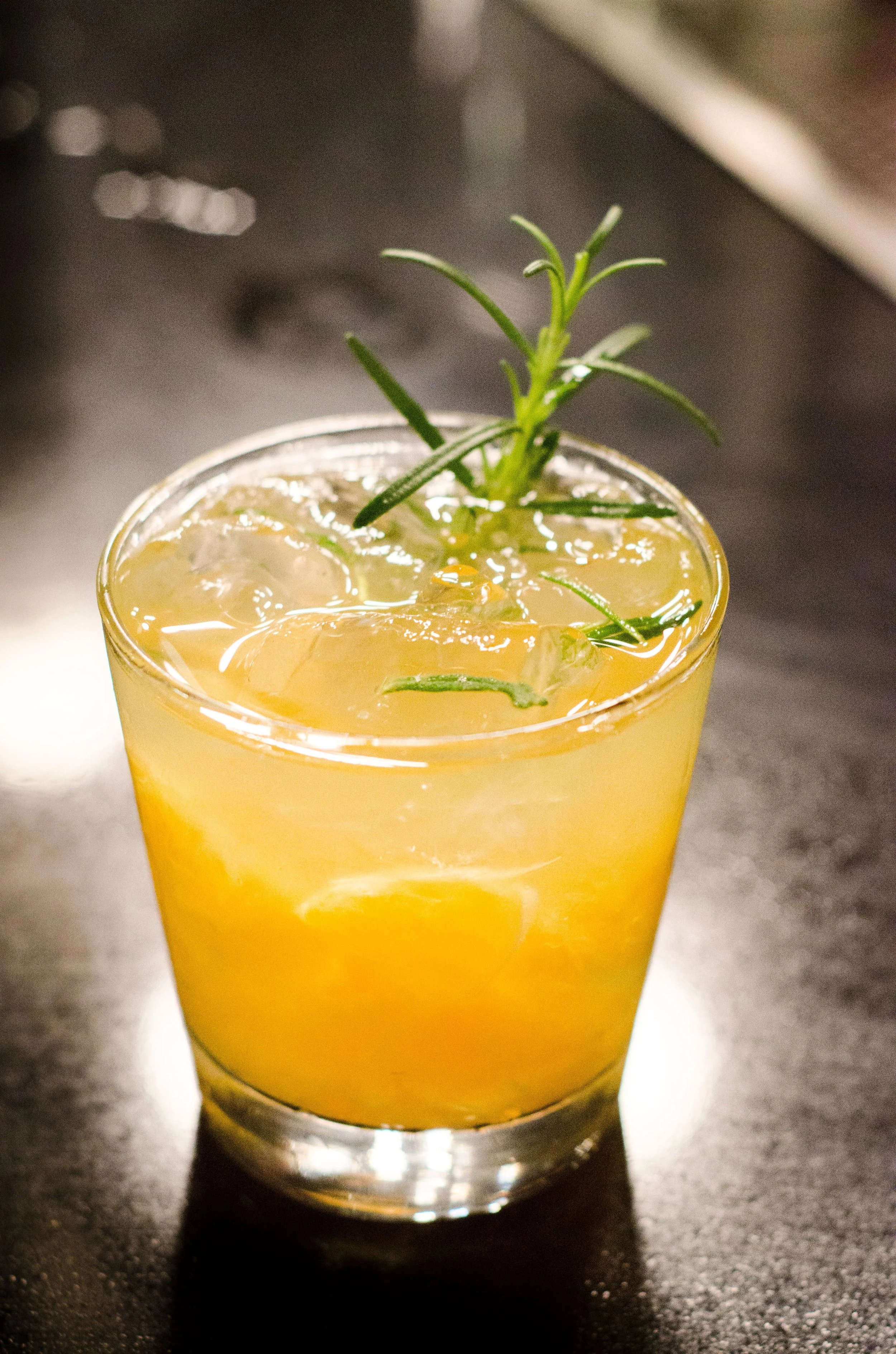 A close-up of a yellow-orange drink in a clear glass filled with ice, garnished with a sprig of green herbs, possibly rosemary.