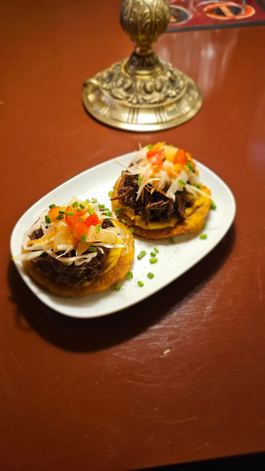 Two Tostones with shredded beef, topped with chopped onions, cilantro, and diced tomatoes, served on a white oval plate. A decorative lamp is in the background on a wooden table.