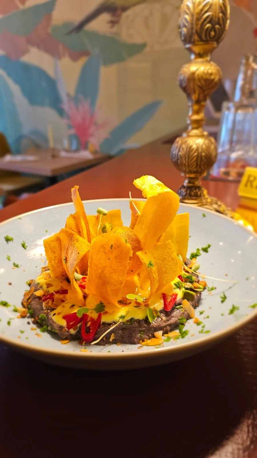 A plated dish featuring plantain chips, garnished with herbs, peppers, and microgreens, set over a dark sauce in a light blue bowl, with a decorative lamp and floral wallpaper in the background.