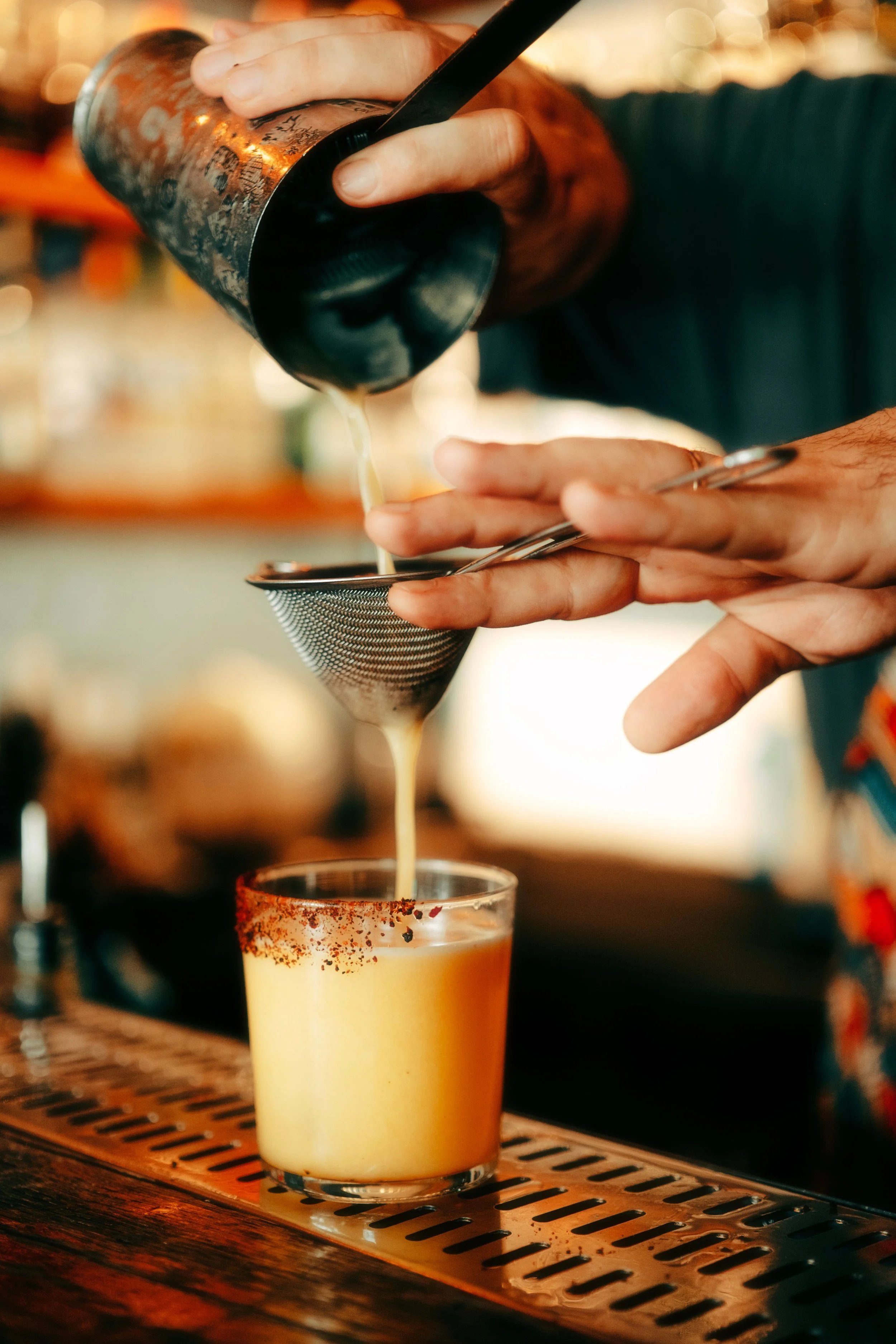 A bartender pours a creamy, yellow-colored drink through a strainer into a glass with a chili powder rim, on a bar counter with a wooden surface.