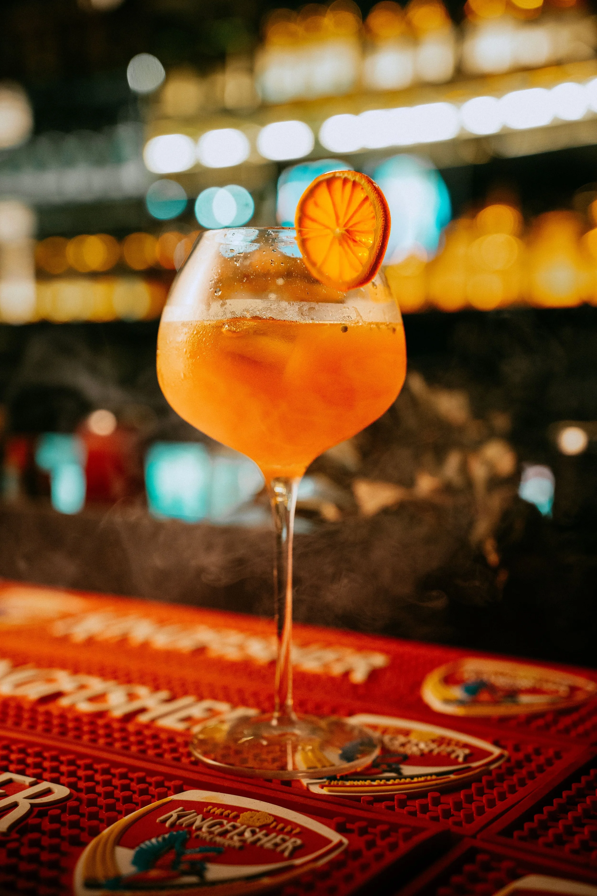 A cocktail glass filled with an orange-colored drink, garnished with a slice of orange or lemon, sitting on a table at a bar or restaurant with blurred colorful lights in the background.