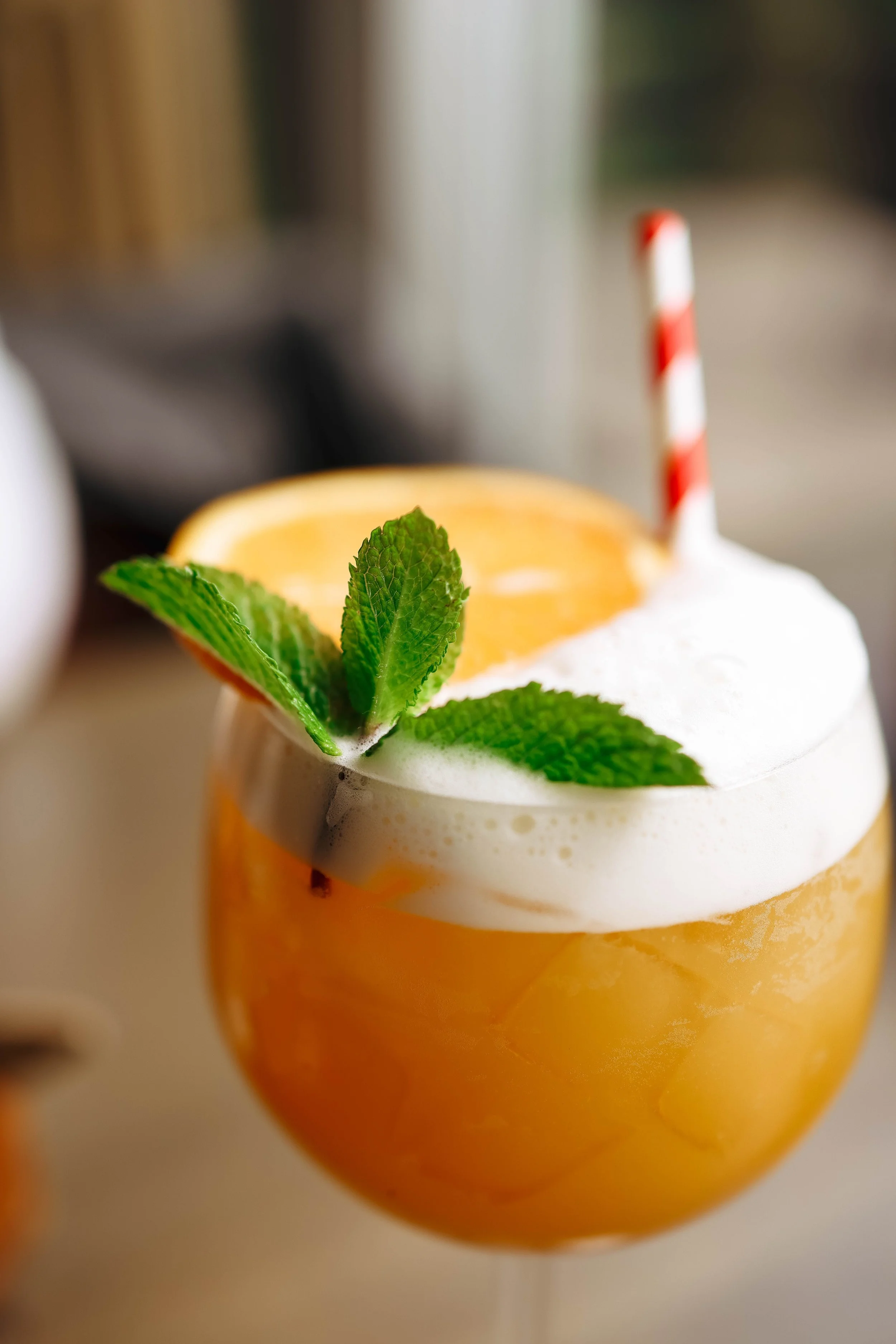 A close-up of a yellow and orange drinking glass garnished with fresh mint leaves and a slice of orange, with a red and white striped straw on a blurred background.