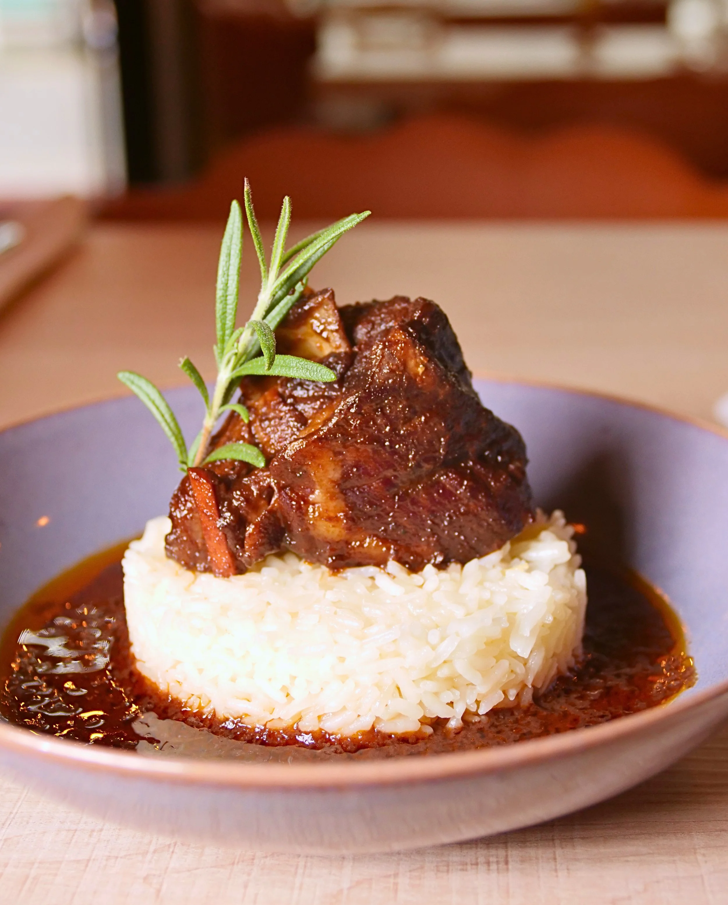 Cooked beef short ribs with brown sauce, garnished with a sprig of rosemary, served on a bed of white rice in a metallic bowl.