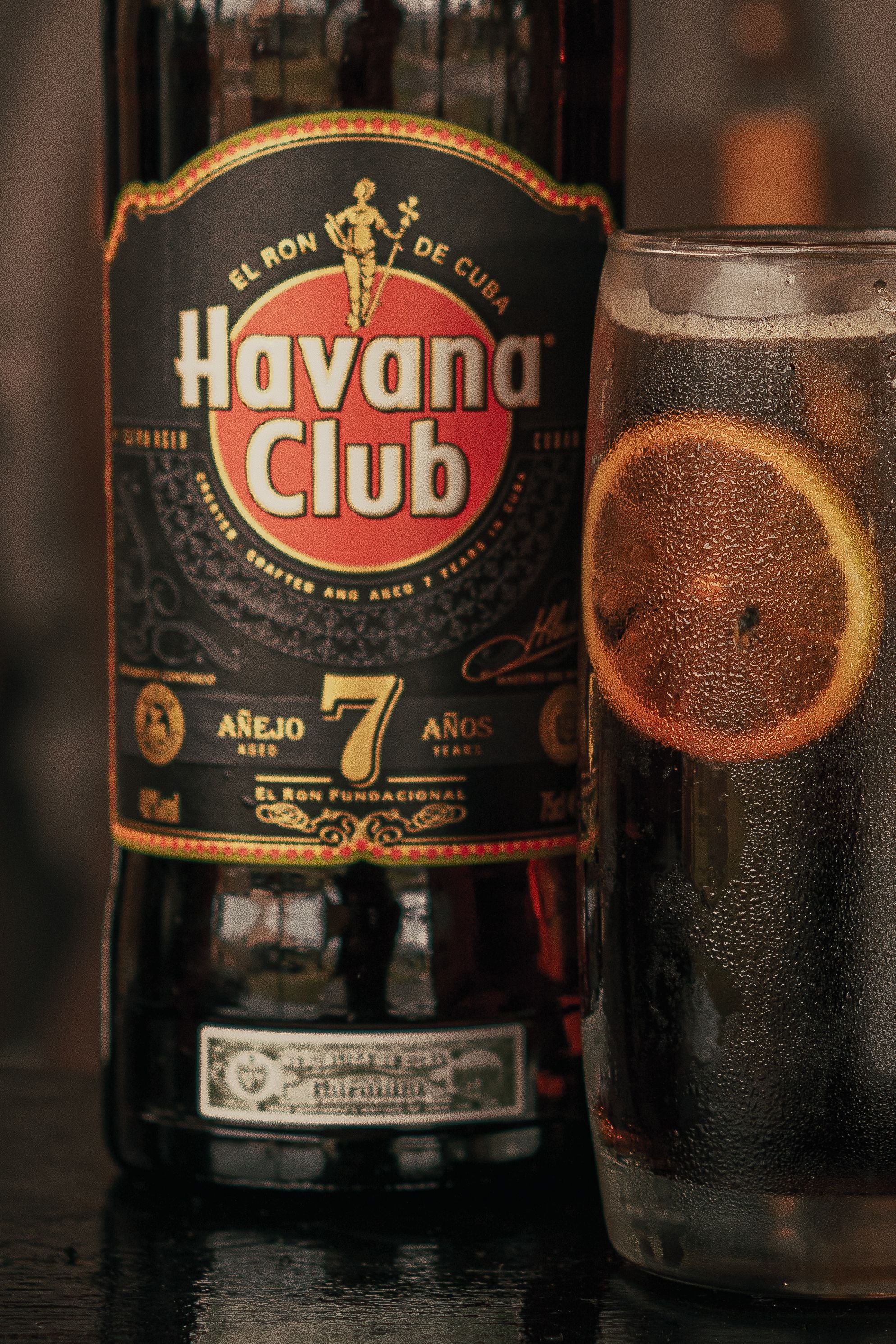 A bottle of Havana Club rum with a label featuring a man in a suit holding a sword and the text "El Ron de Cuba." Next to the bottle is a glass of soda with ice and lemon slices.