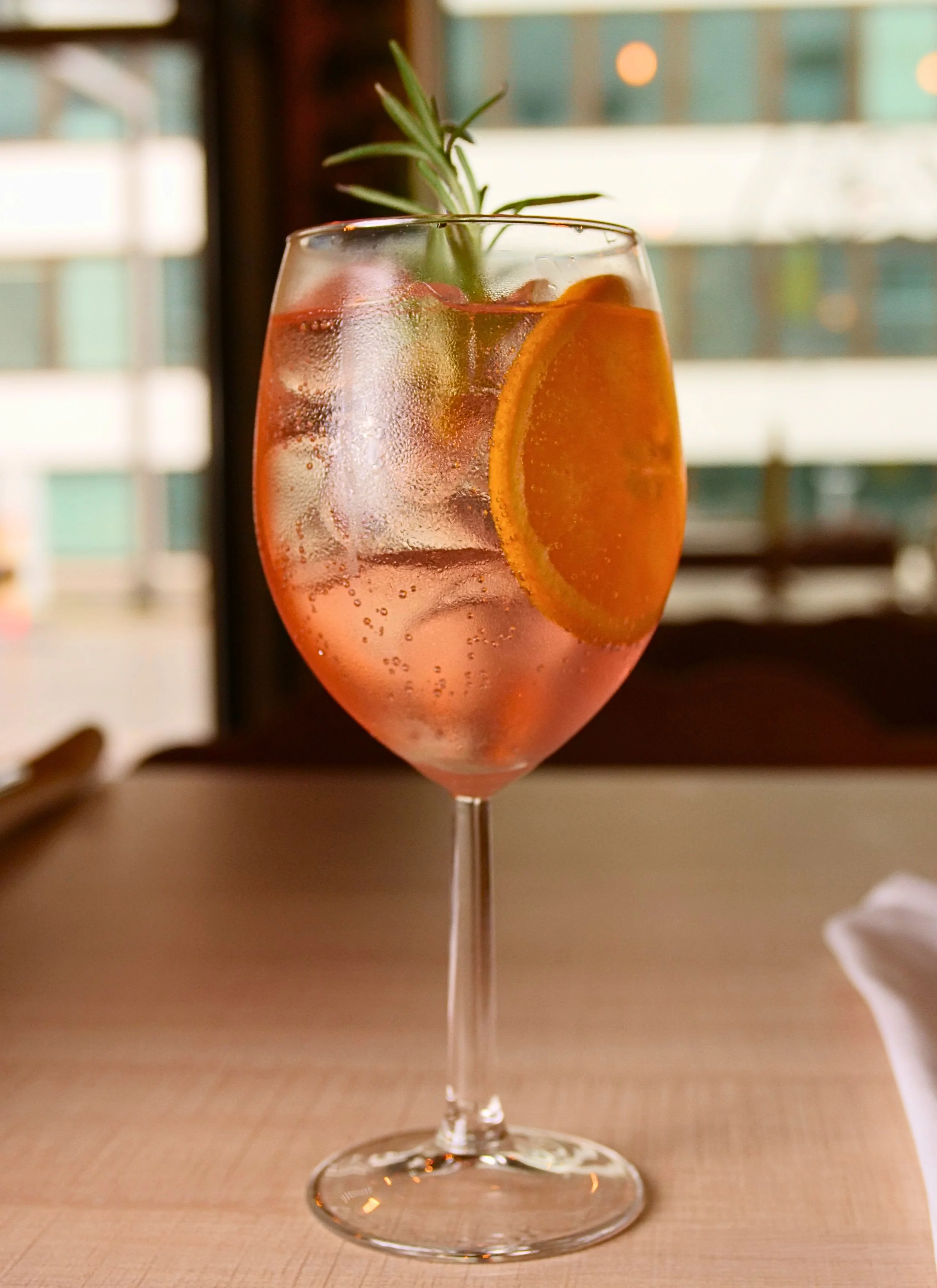 A glass of pink cocktail with ice, a slice of orange, and a sprig of rosemary on top, placed on a wooden surface.