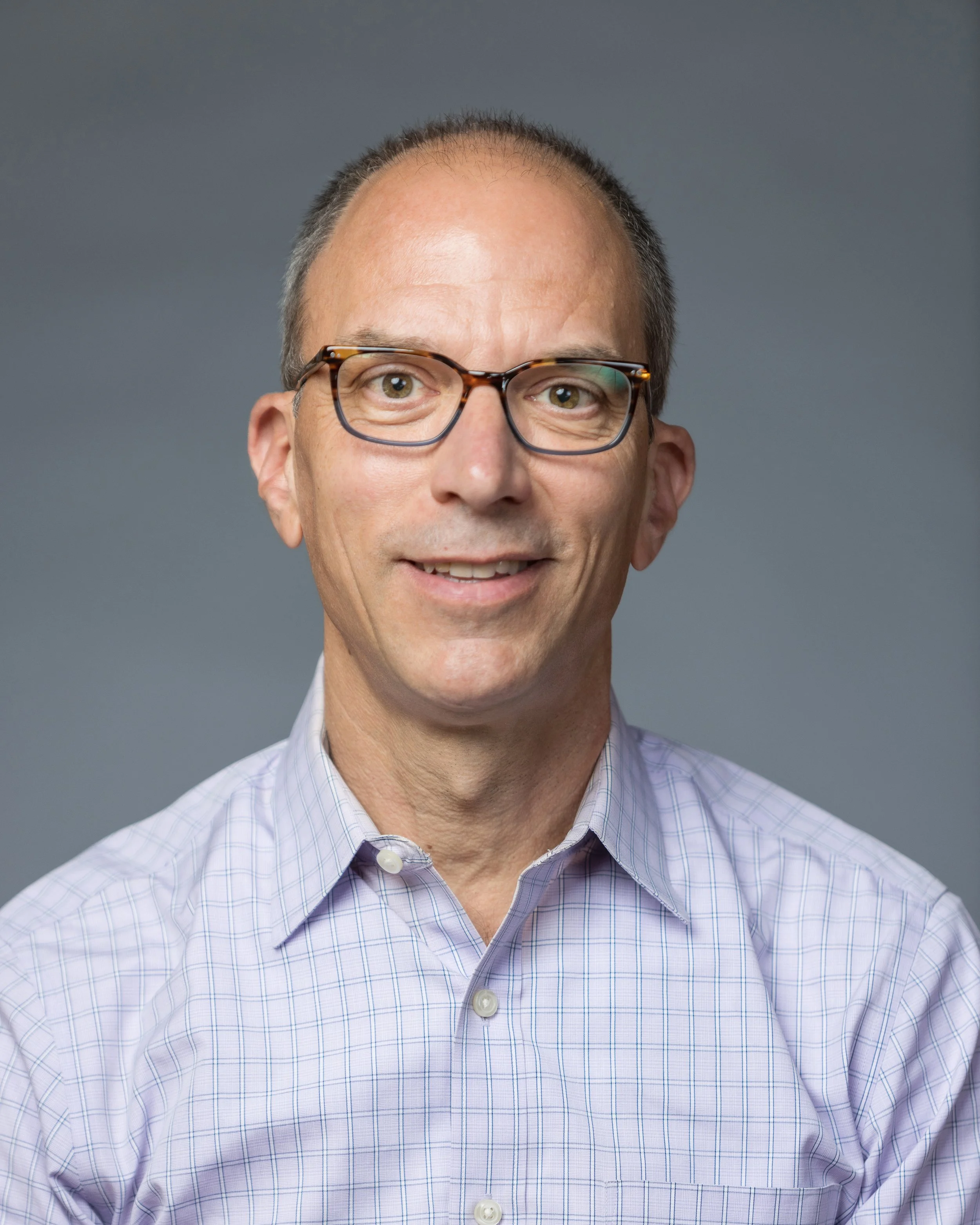 A middle-aged man with short hair, wearing tortoiseshell glasses and a light purple, checkered dress shirt, smiling against a plain, gray background.