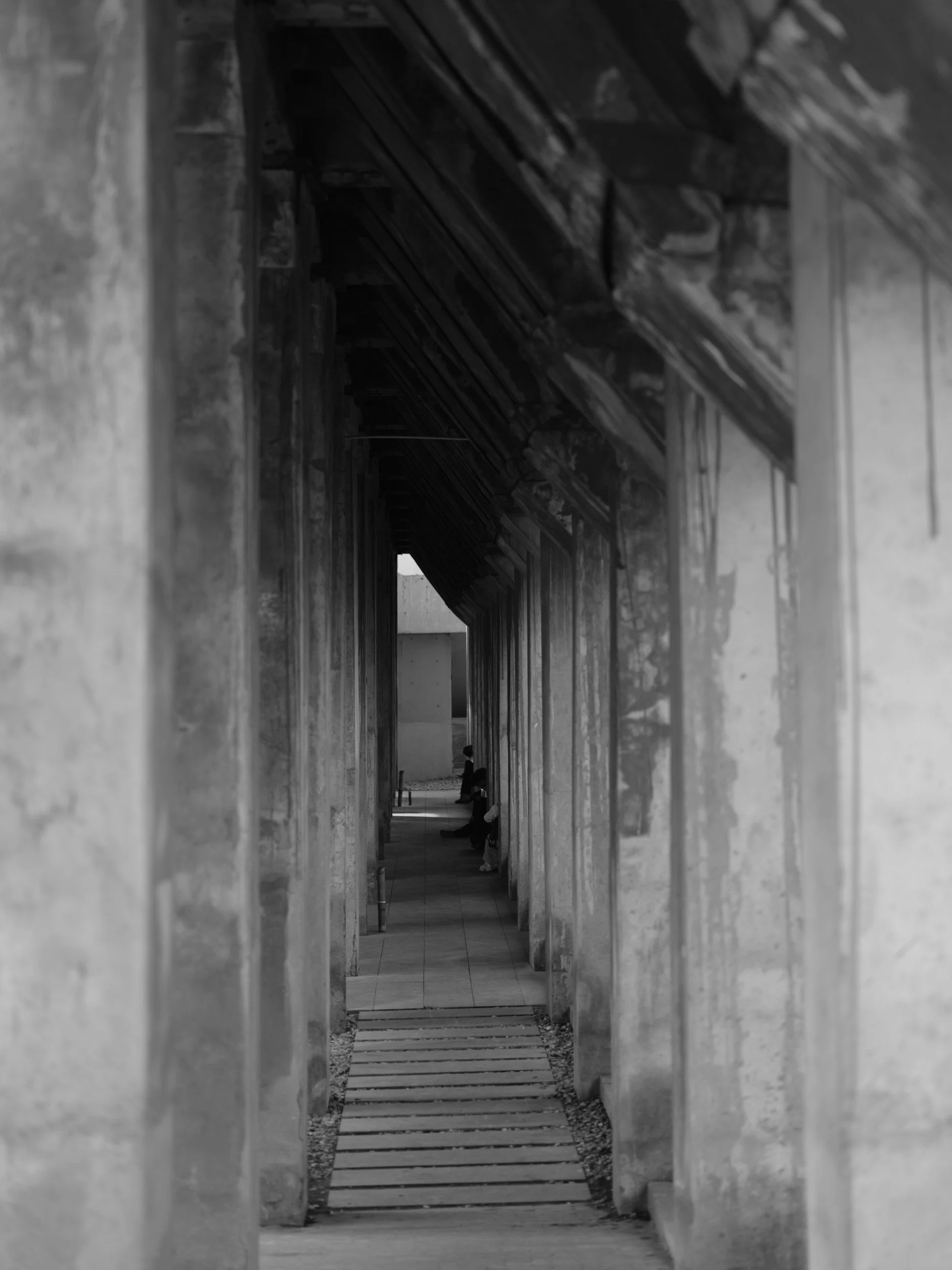 Black and white photo of a narrow exterior corridor with concrete walls on either side, leading to an archway with a hallway and two seated individuals at the far end.