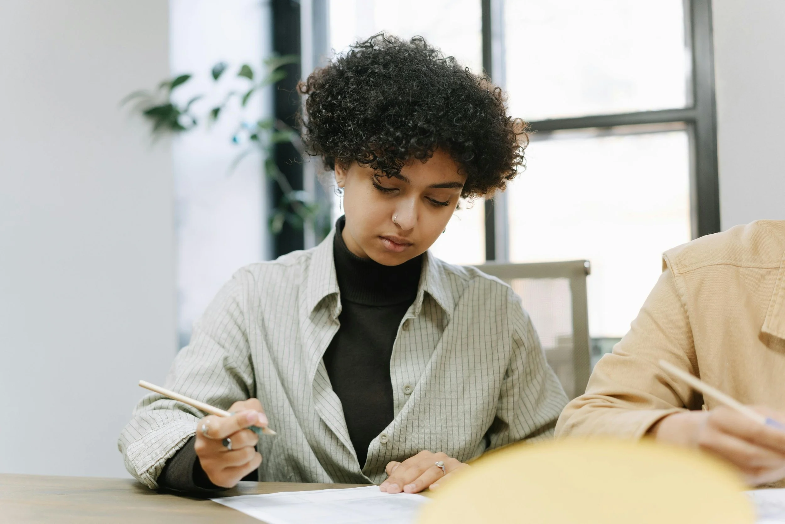 A young woman with curly hair, wearing a striped shirt and black turtleneck, writing with a pen at a desk in a bright office space.