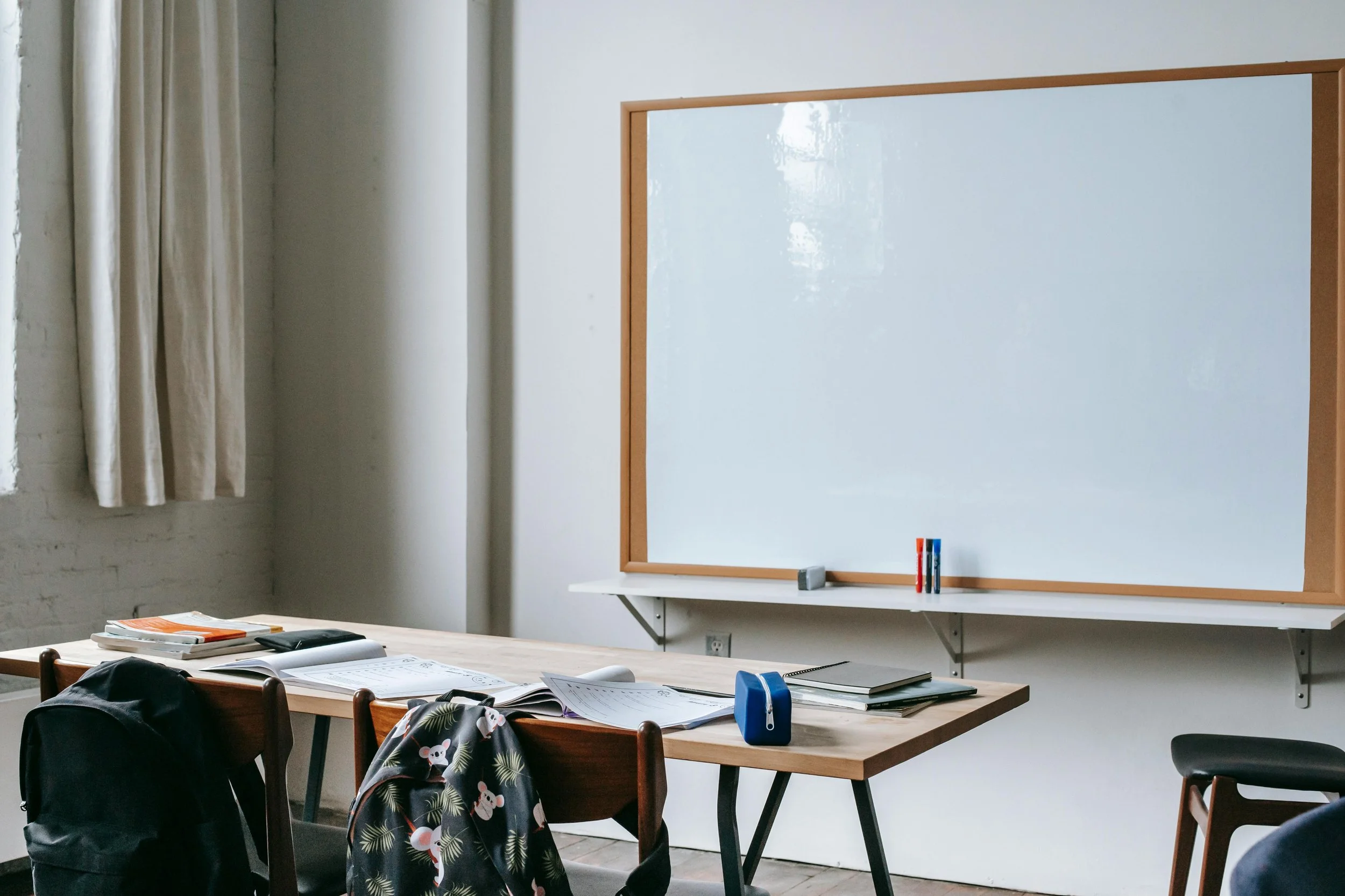 An empty classroom with a whiteboard on the wall, a table cluttered with papers and books, chairs, and a backpack. There are curtains on the window and some writing supplies on the whiteboard.
