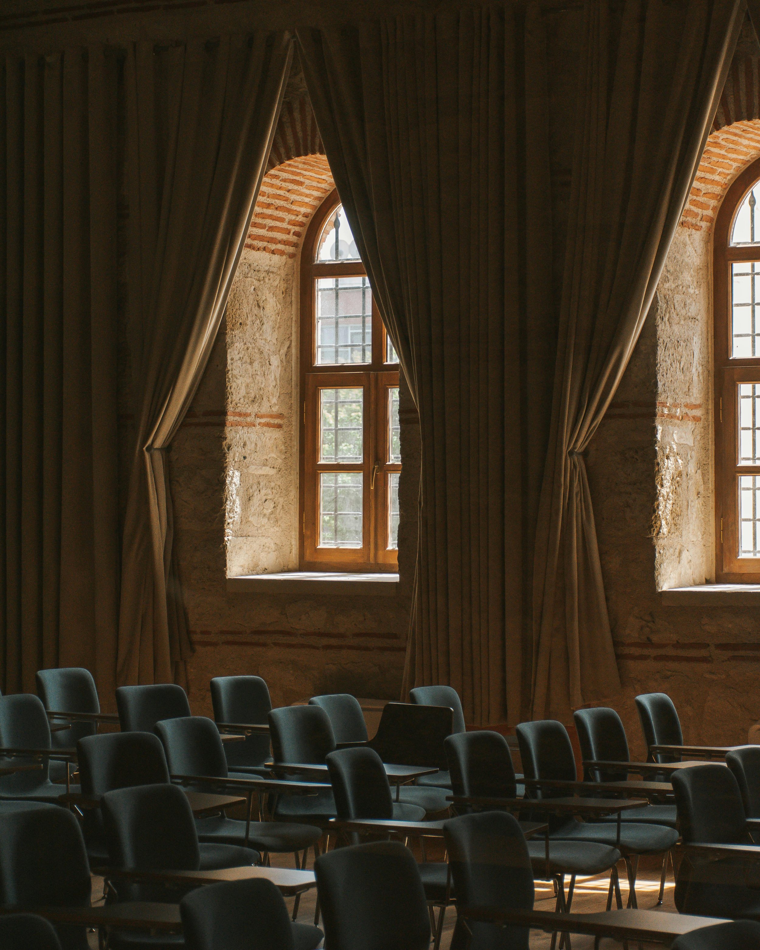 Interior of a room with large arched windows, brick walls, heavy brown curtains, and black chairs arranged in rows.