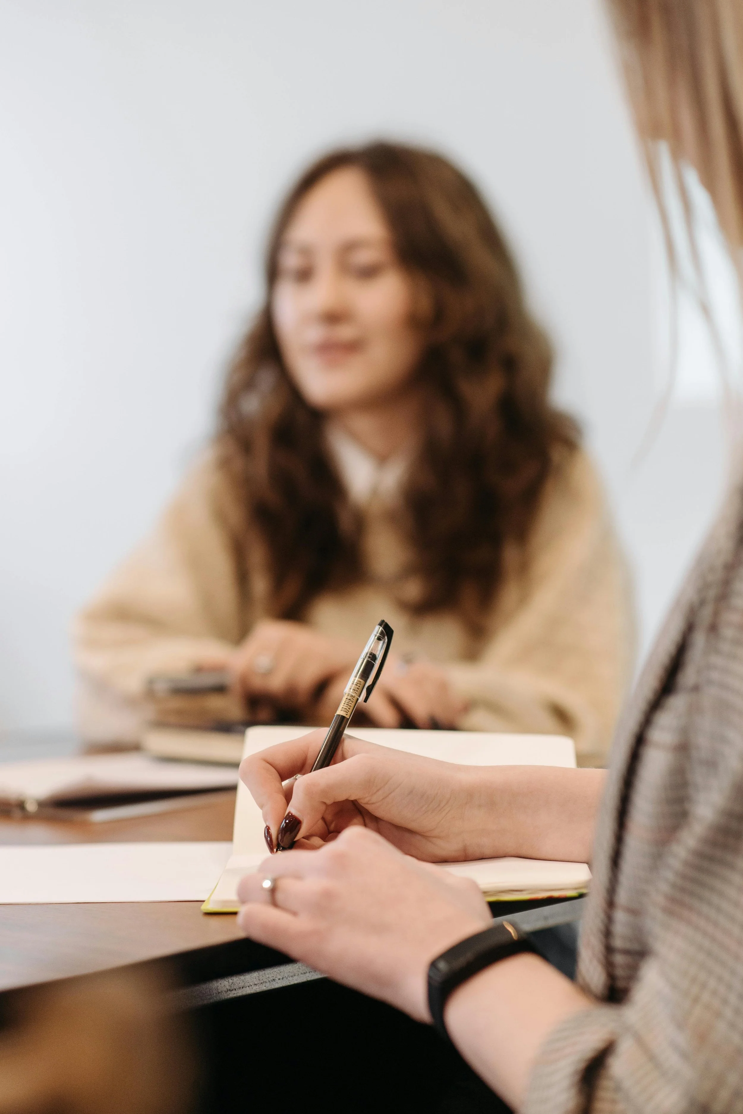 A woman with brown hair during a meeting or interview, in focus, with another person's hands holding a pen and notebook in the foreground, and a woman with curly brown hair in the background.