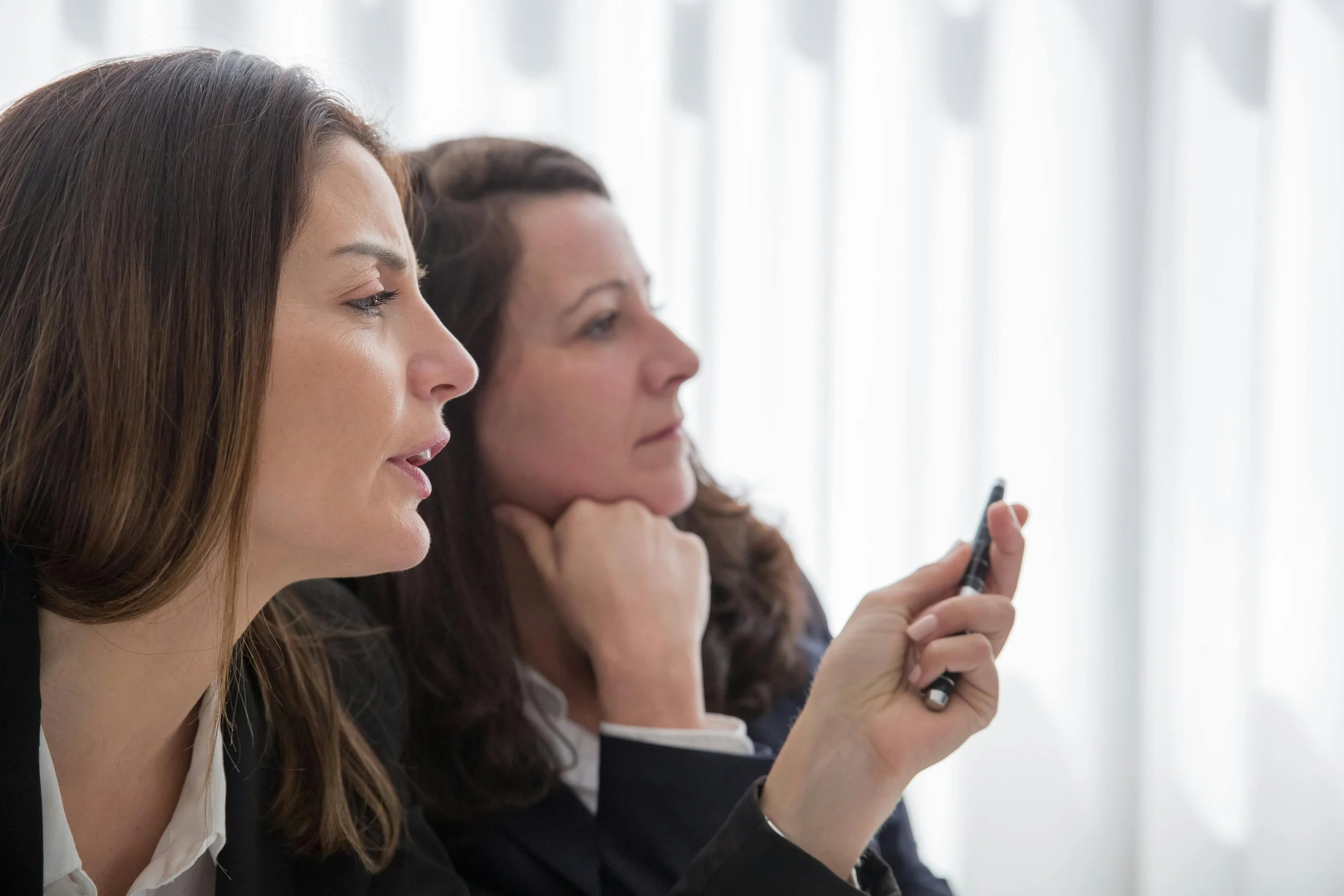 Two women sitting side by side, one with long brown hair and the other with curly brown hair, looking at a smartphone held by the woman with curly hair in a bright room with vertical blinds.