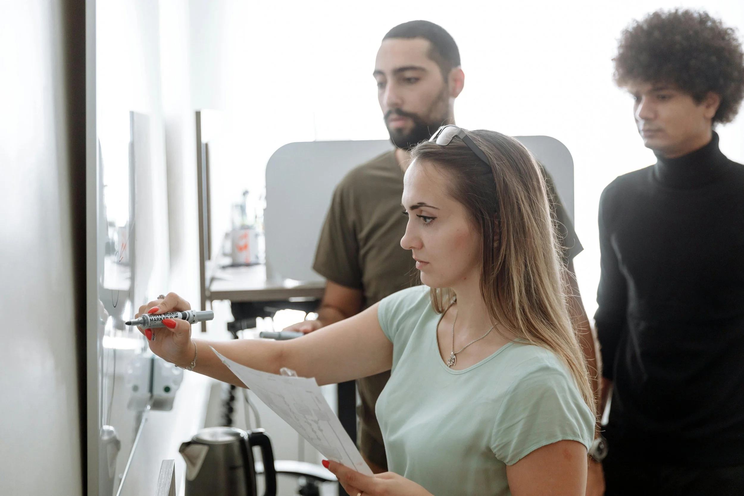 A woman is writing on a whiteboard with a marker, while two men observe her in a bright room.