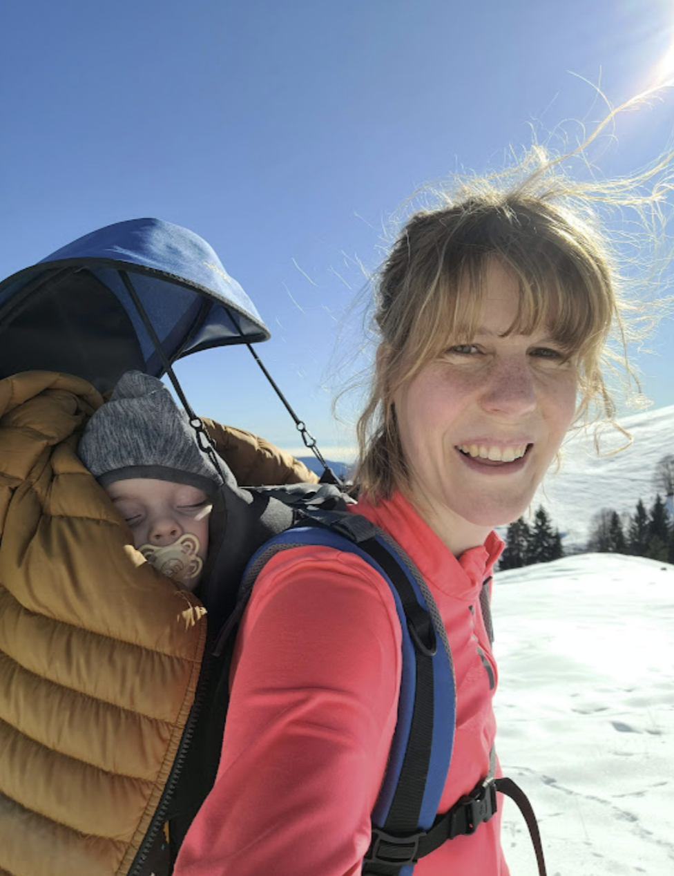 A woman smiling as she carries a sleeping baby in a backpack carrier on a snowy mountain with clear blue sky and bright sunlight.