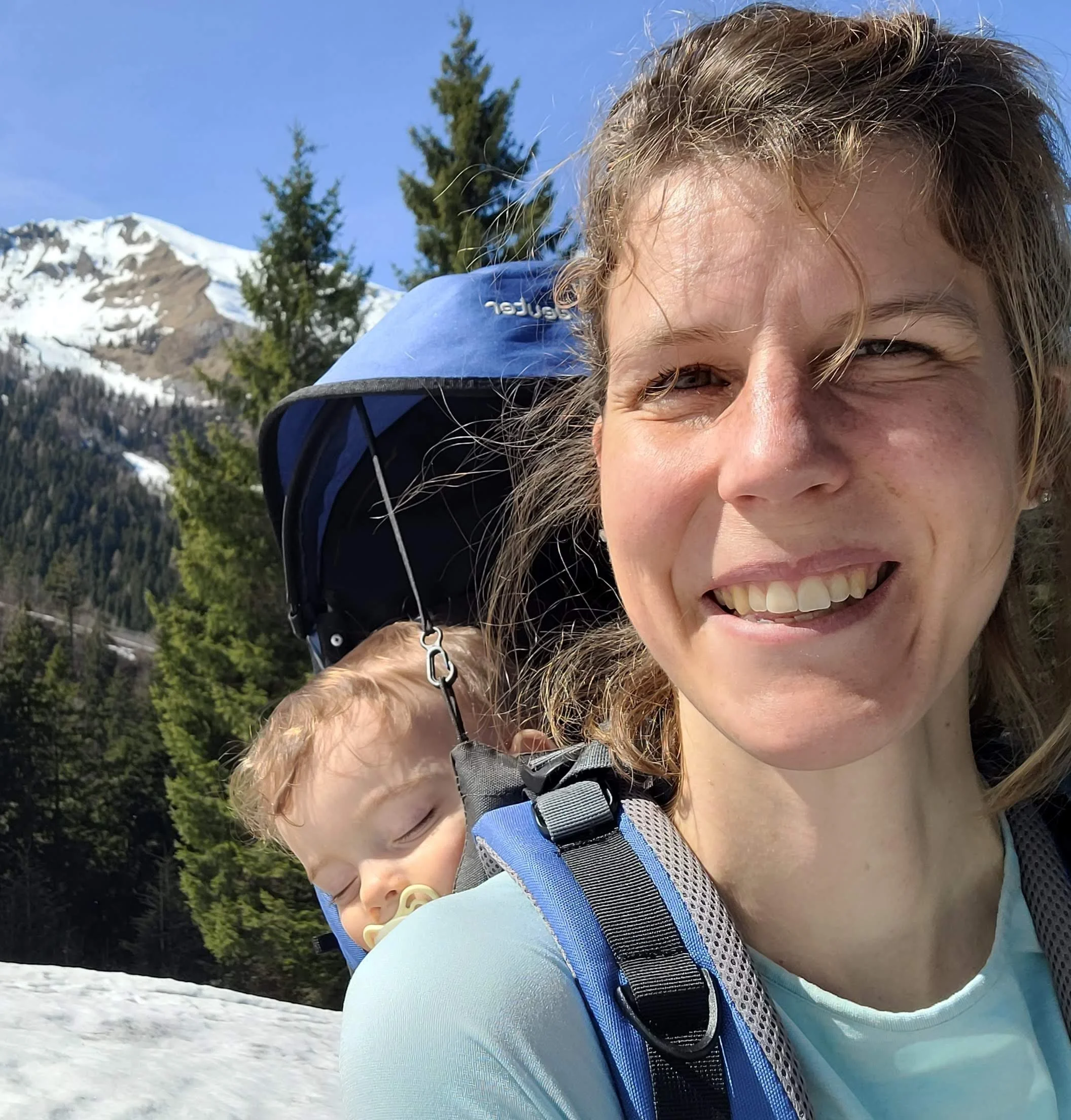 Smiling woman with a backpack outdoors in a snowy mountain landscape with trees and snow-covered peaks.