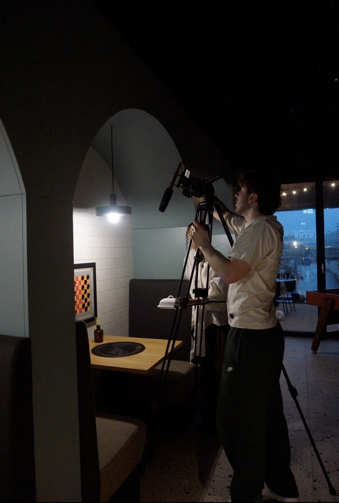 Two people filming a video in a dimly lit indoor space during evening, with a large camera on a tripod aimed at a wall with a framed artwork.