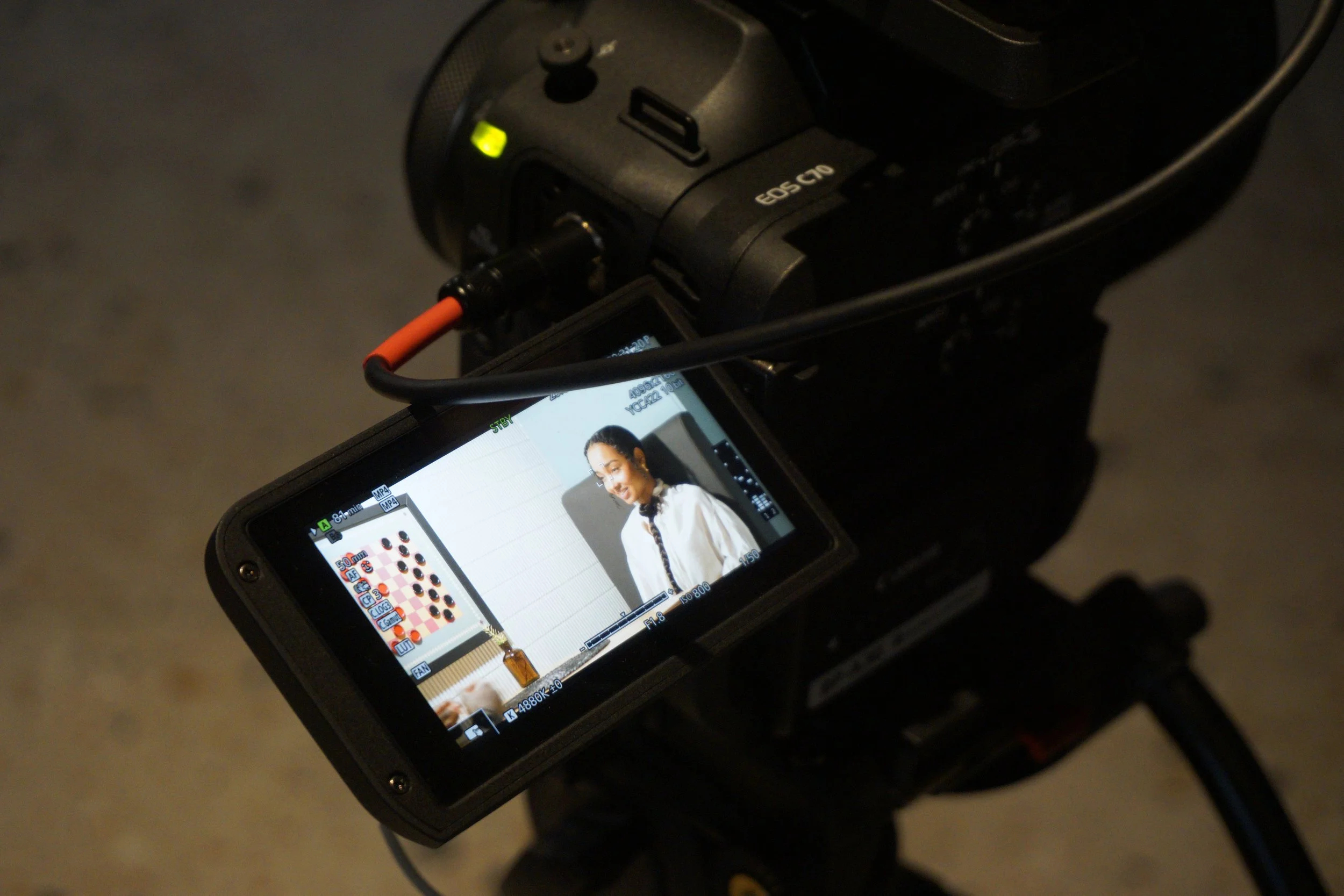 A digital camera's screen displaying an image of a woman with dark hair, wearing a white blouse, in front of a white background. The camera is connected to a device with an orange cable, and there is a small green light on the camera. The background of the photo is dark and blurred.