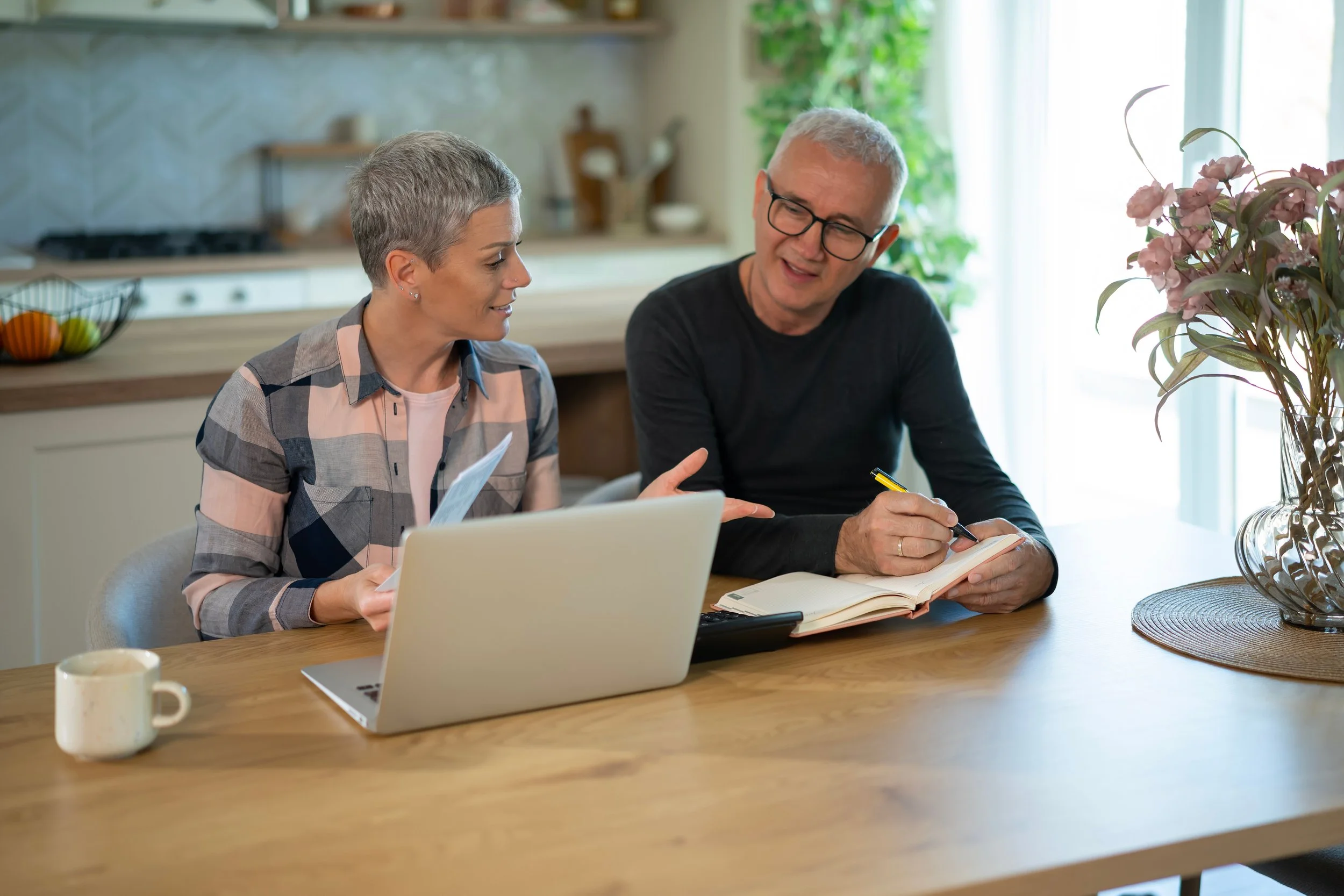 A man and woman sitting at a wooden table in a bright kitchen. They are smiling and engaged in conversation, with a laptop, notebooks, and a pen on the table. A large vase with pink flowers is on the right side of the table.