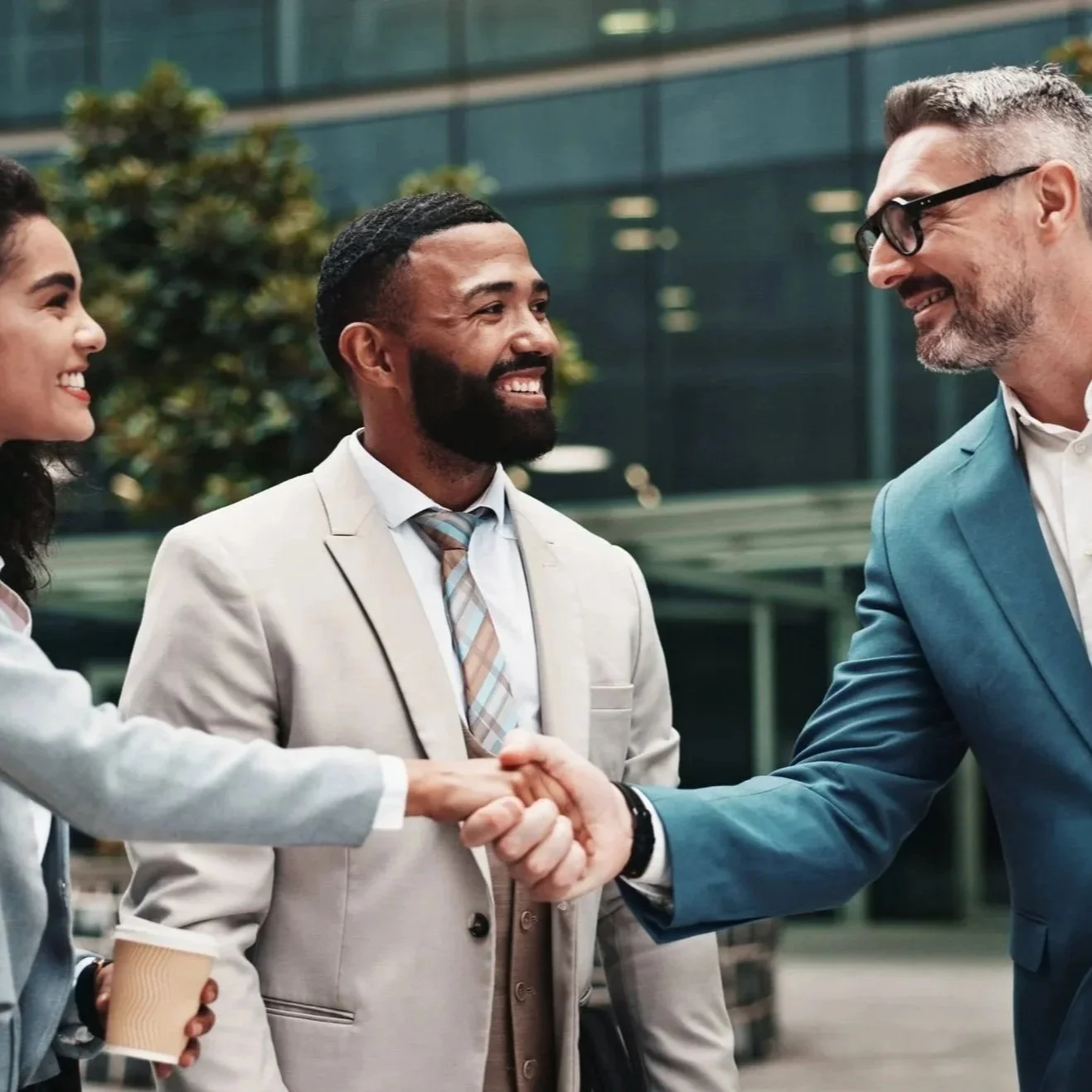 Three business professionals, two men and a woman, smiling and shaking hands outdoors in front of a modern office building.