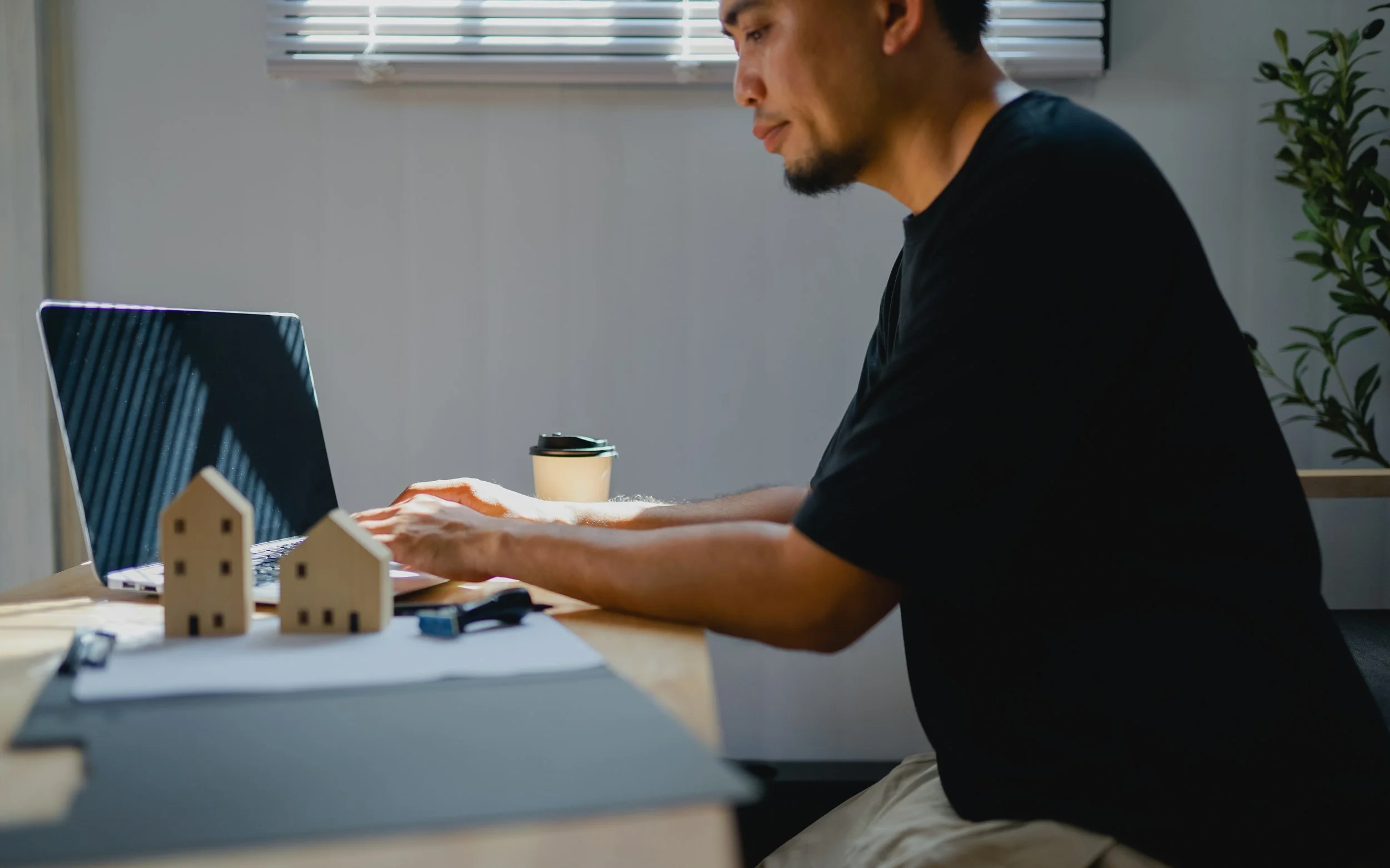 A man working on a laptop at a desk with small wooden house models scattered around, a coffee cup nearby, and a plant in the background.