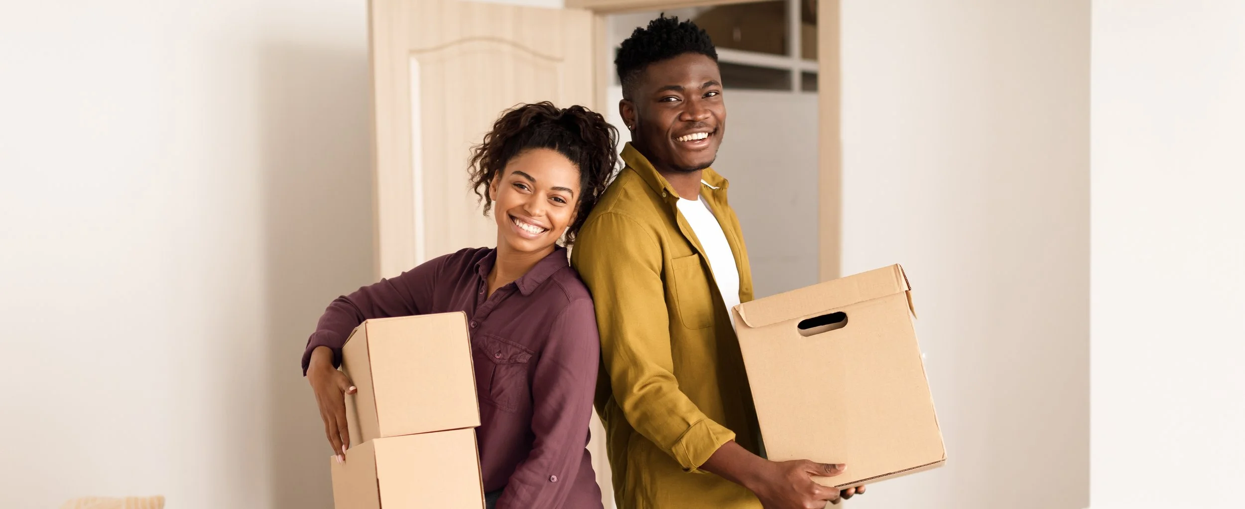 A young woman and young man are standing next to each other, smiling, each holding cardboard boxes. They are inside a house with light-colored walls, possibly moving in or out.
