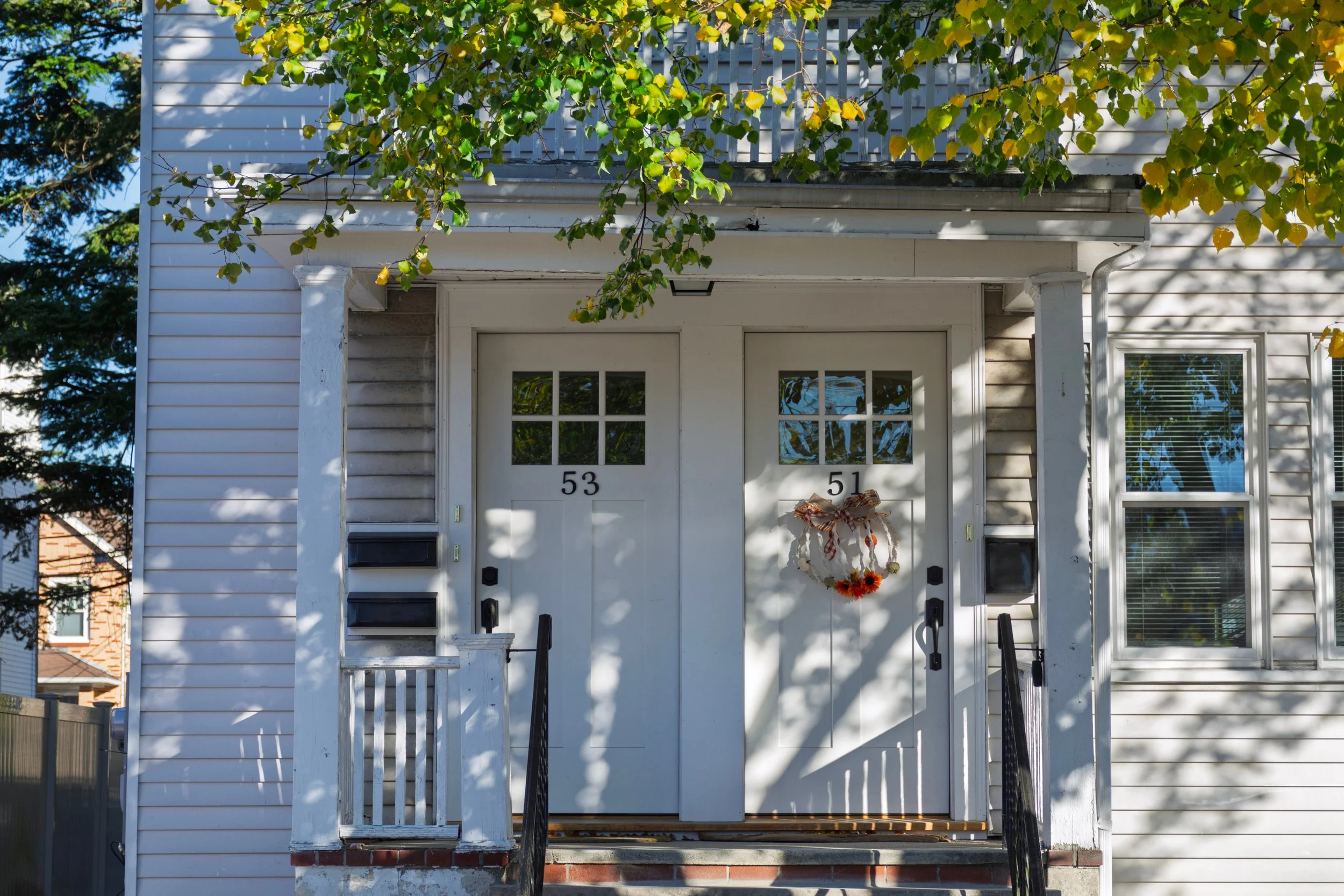 Front view of a white house with two front doors labeled 51 and 53, decorated with a fall-themed wreath, and surrounded by green and yellow leaves, casting shadows on the facade.