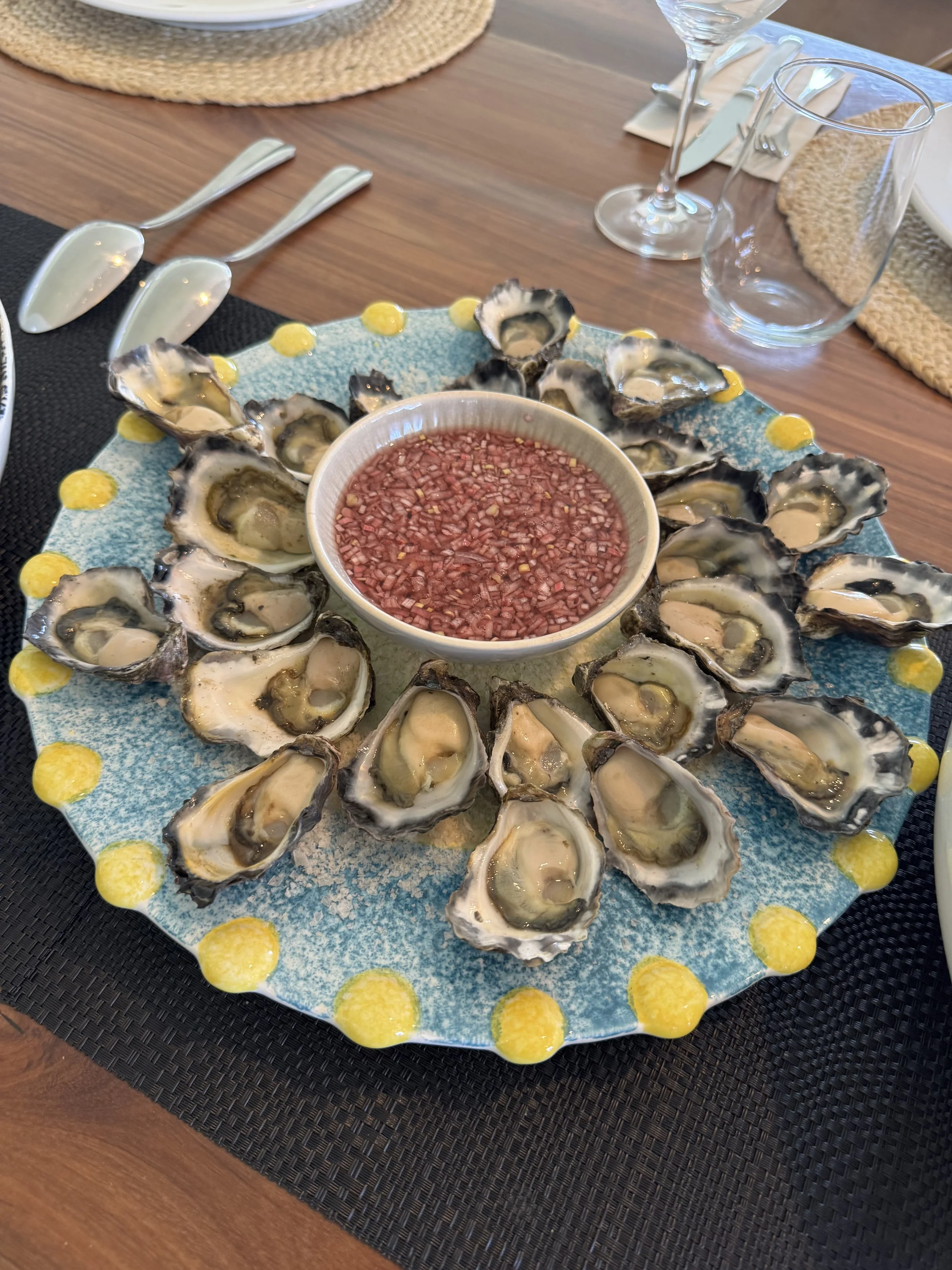 A plate of fresh oysters on the half shell arranged in a circle with a bowl of cocktail sauce in the center, set on a table with glassware and utensils.