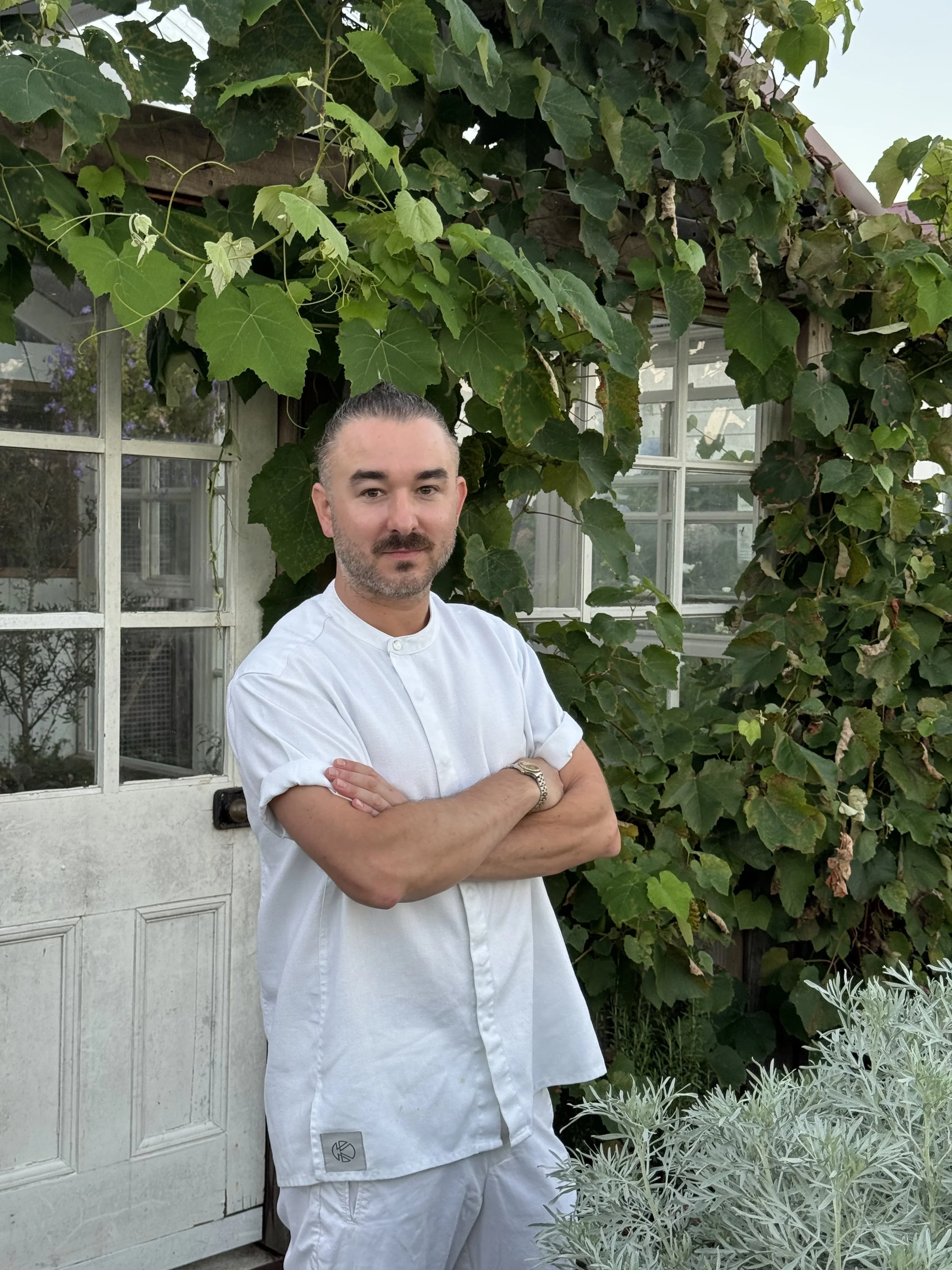 A man standing outdoors with arms crossed, wearing a white chef's coat and pants, in front of a leafy green vine-covered structure with windows.