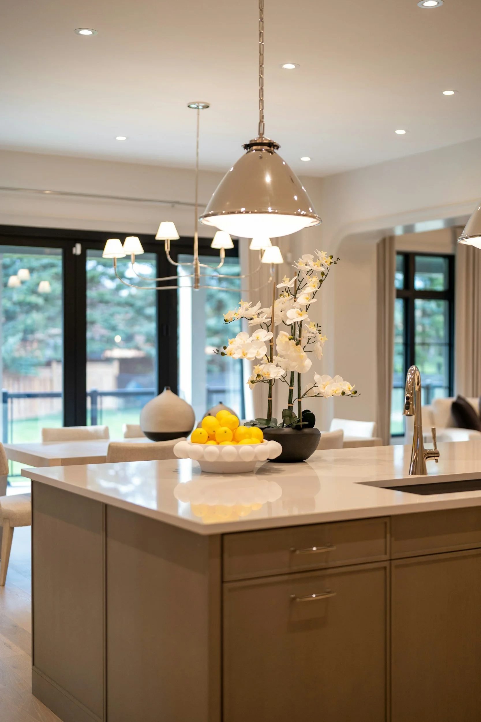 Kitchen island with potted white orchids, a bowl of yellow lemons, a decorative vase, and a kitchen sink, with large windows and family room in background.