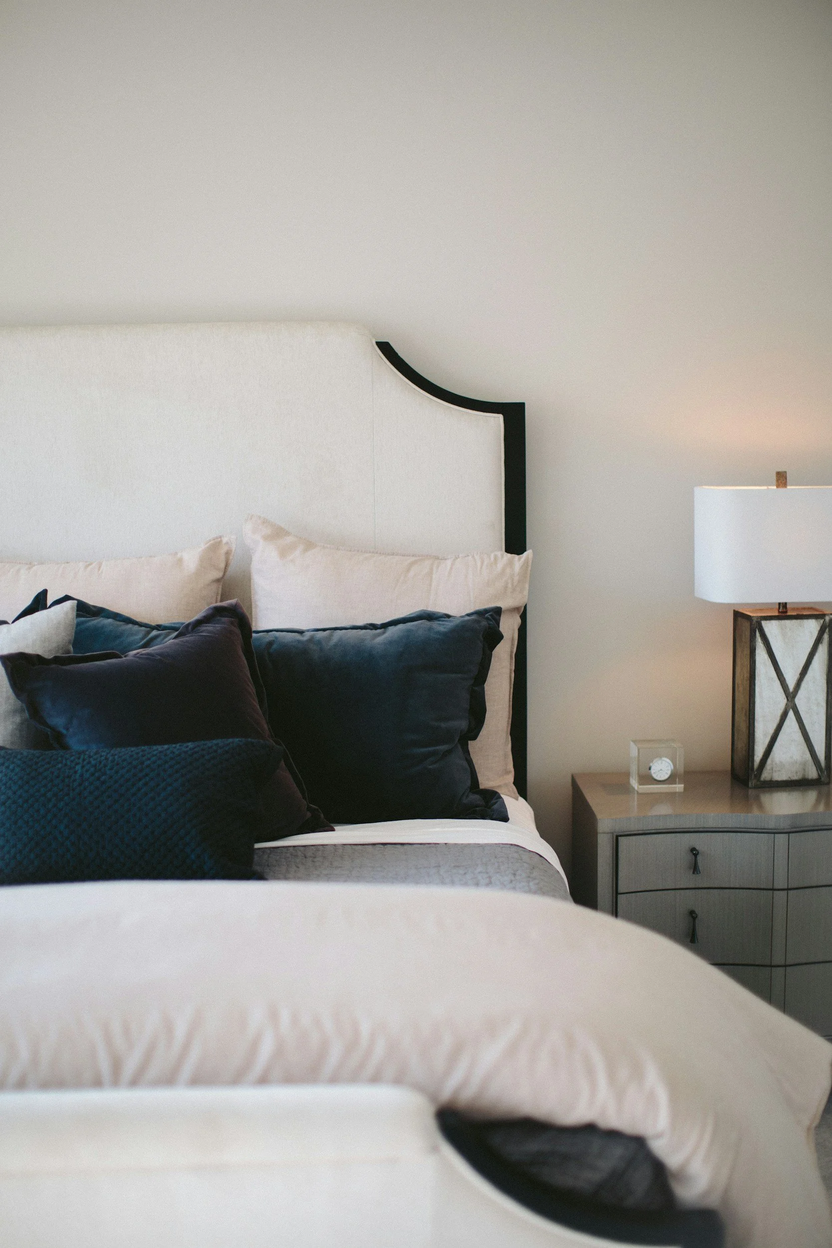 Close-up of a bed with dark and light pillows, a nightstand with a small clock, a glass of water, and a framed lamp beside it, in a bedroom with a neutral wall color and minimal decor.