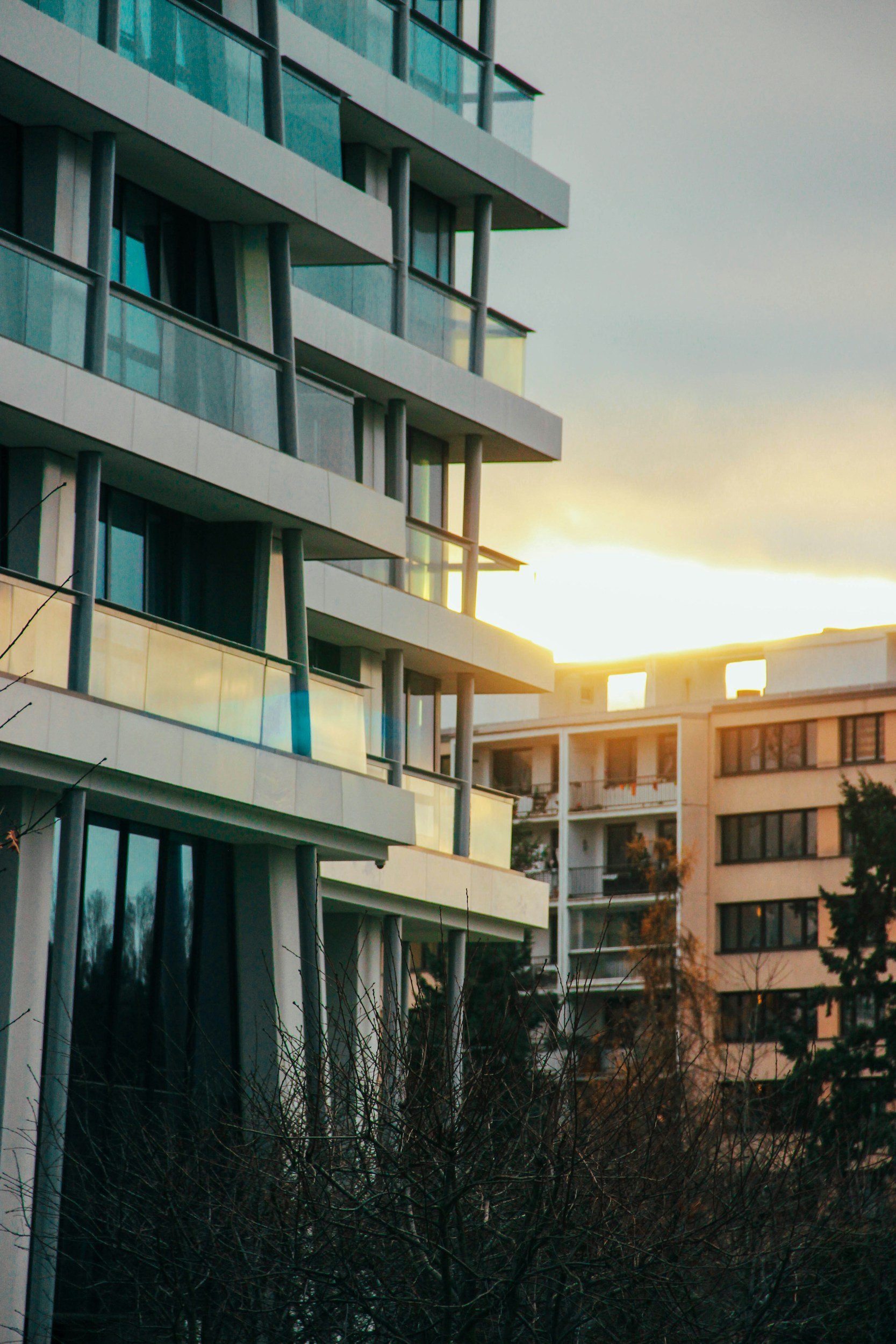 Modern multi-story residential building with glass balconies, set against a sunset sky, with trees and another building in the background.