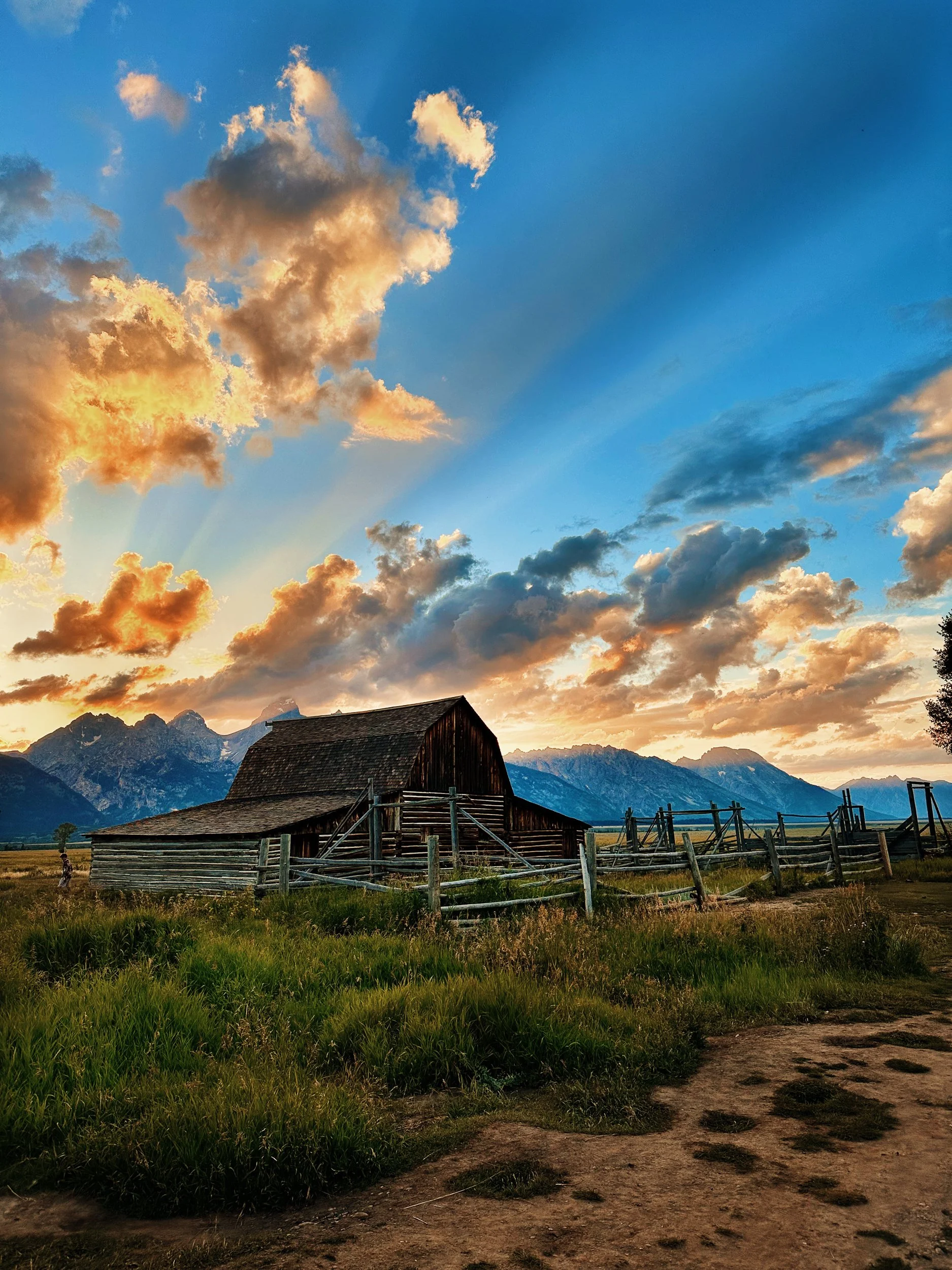 A rustic wooden barn with a sloped roof in a grassy field with a dirt path. There are mountains in the background under a sky filled with clouds during sunset.