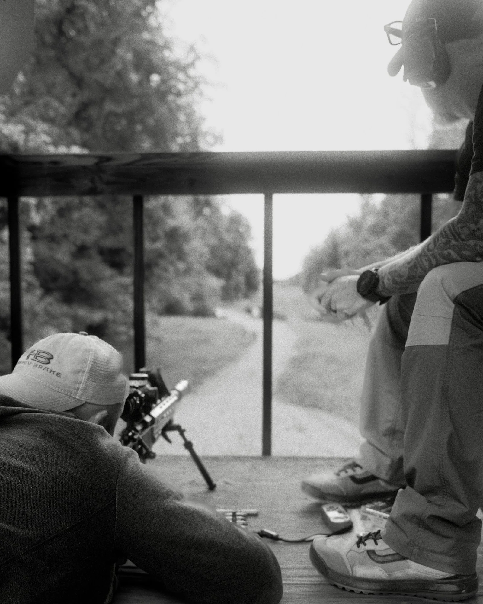 A person wearing sneakers and cargo shorts sitting on the ground with their legs crossed, holding a rifle or sniper scope, with another person wearing a baseball cap and a hoodie sitting nearby, both outdoors near a fence and trees in the background.