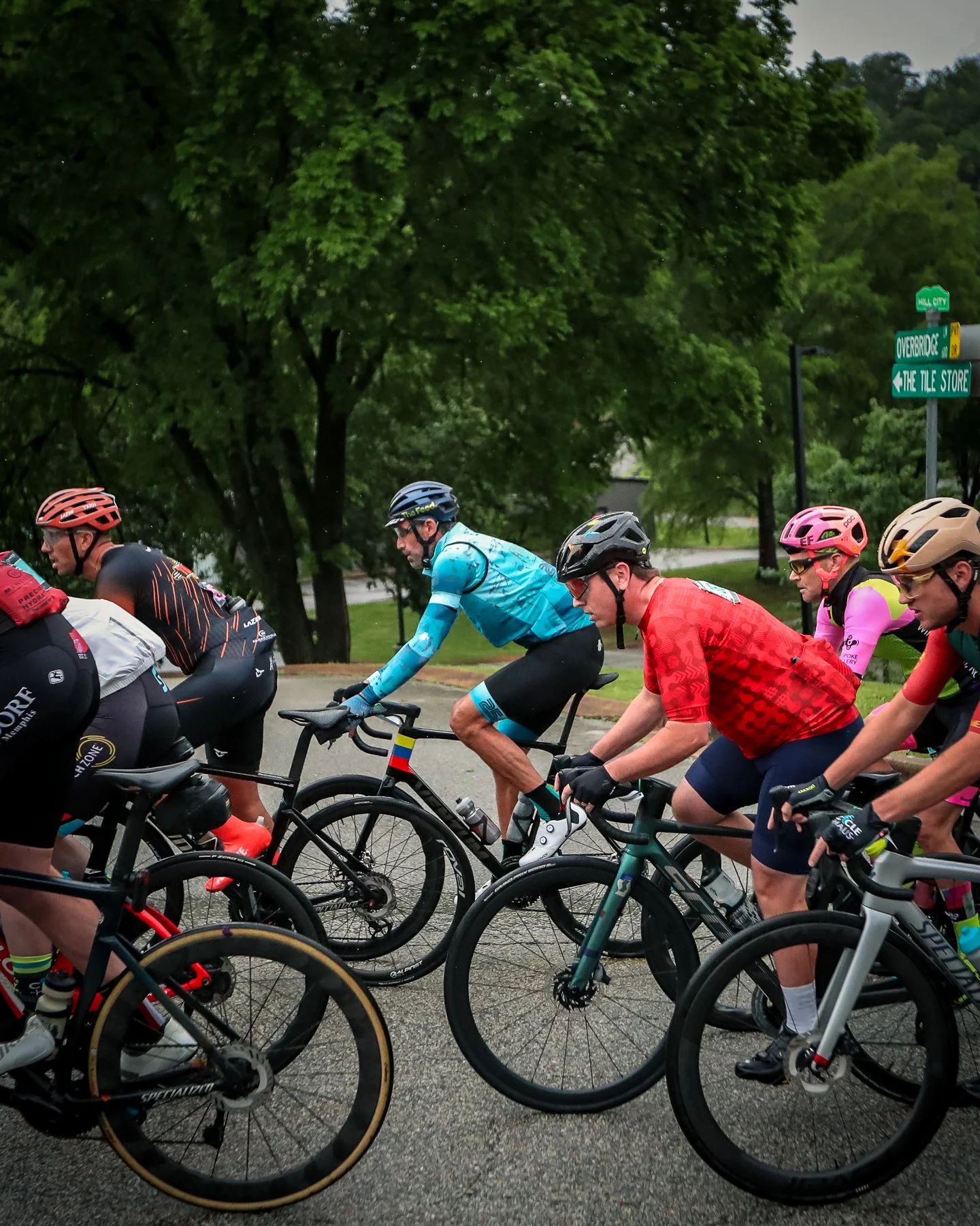 A group of cyclists riding together on a street, with green trees and street signs in the background.