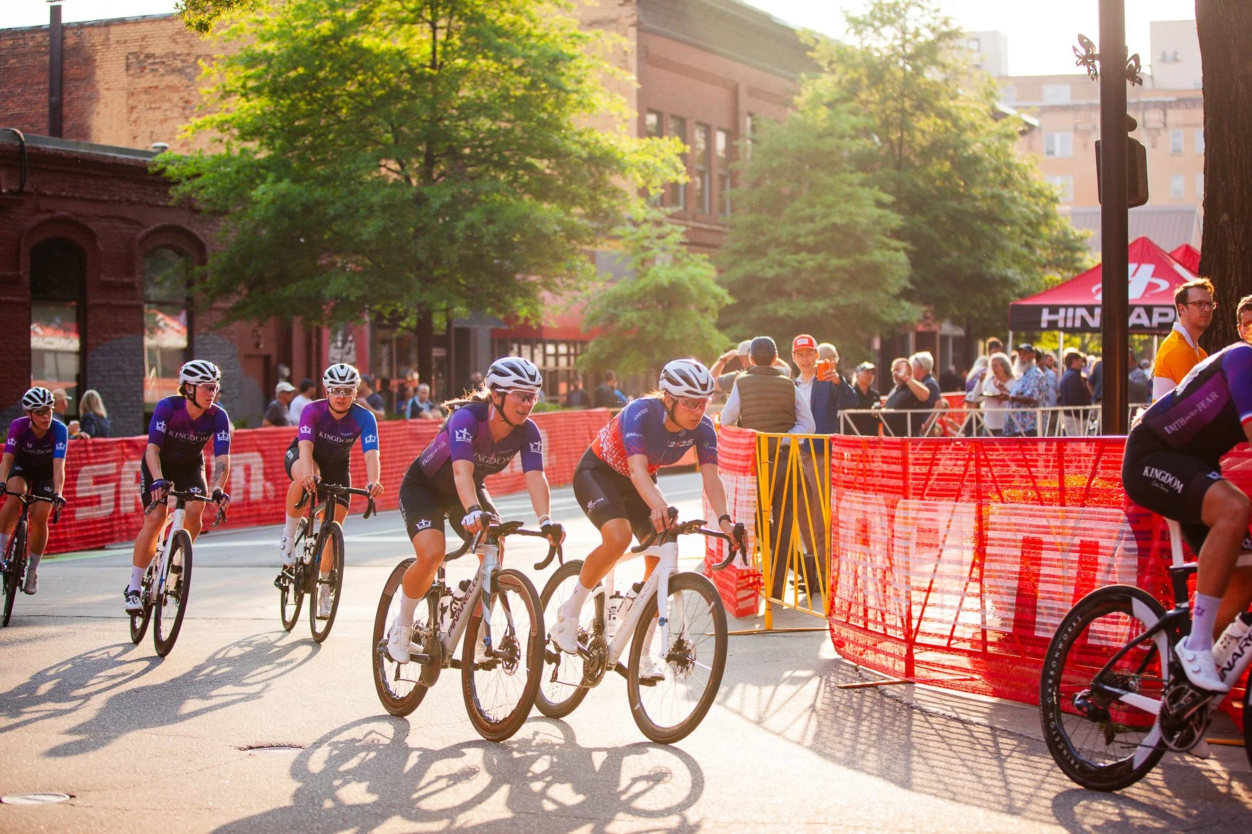 Cyclists participate in a city race on a closed street, with spectators watching behind safety barriers and a canopy in the background.