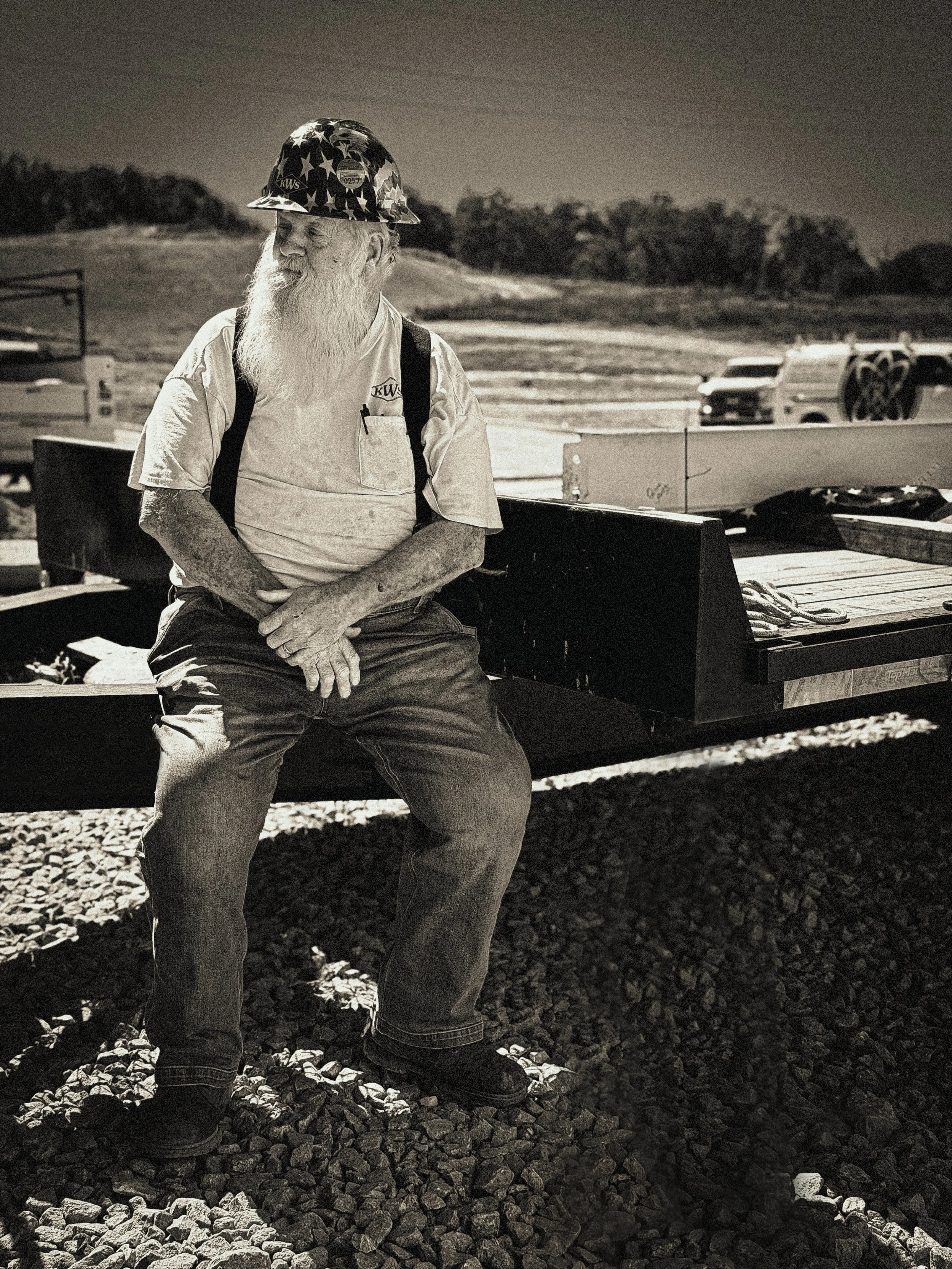 An elderly man with a long white beard wearing a helmet decorated with American flags, a work shirt with a logo, and jeans, standing beside a flatbed trailer on rocky ground at night.